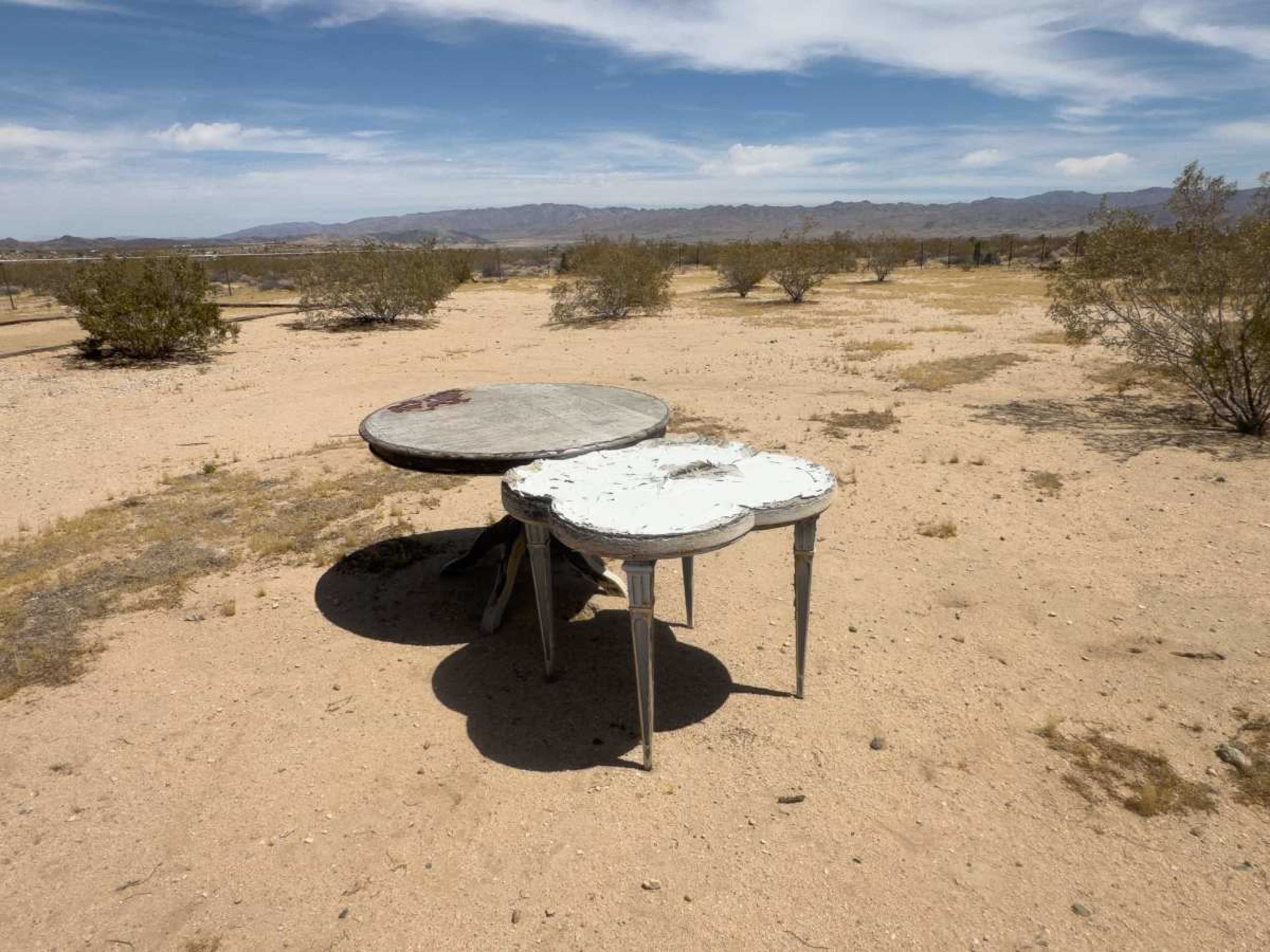 Two weathered tables, one round and one oval, stand in a barren desert landscape surrounded by sparse shrubs and distant mountains.