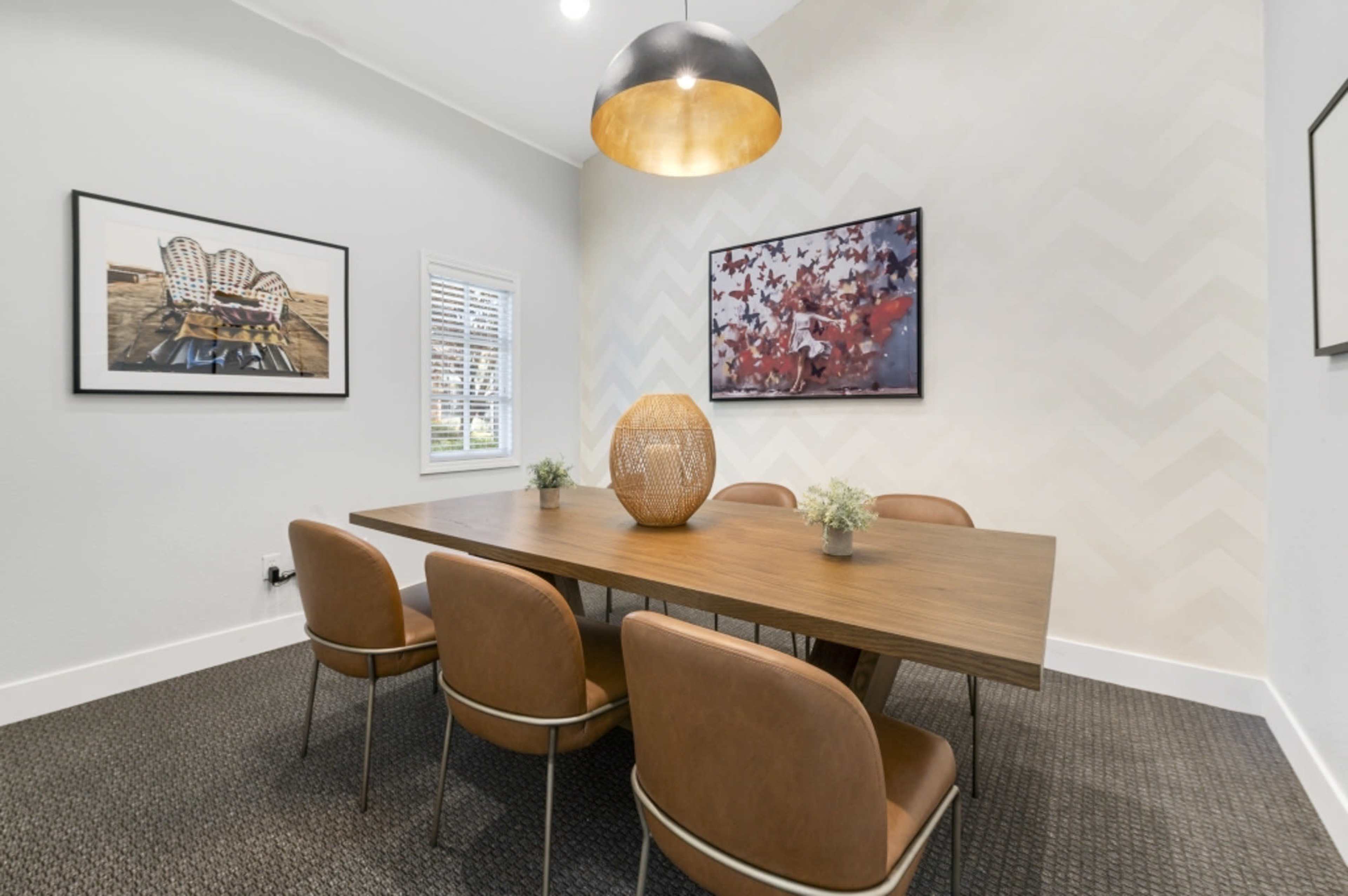 A modern dining room with a wooden table surrounded by six brown chairs, featuring artwork on the walls and a pendant light overhead.
