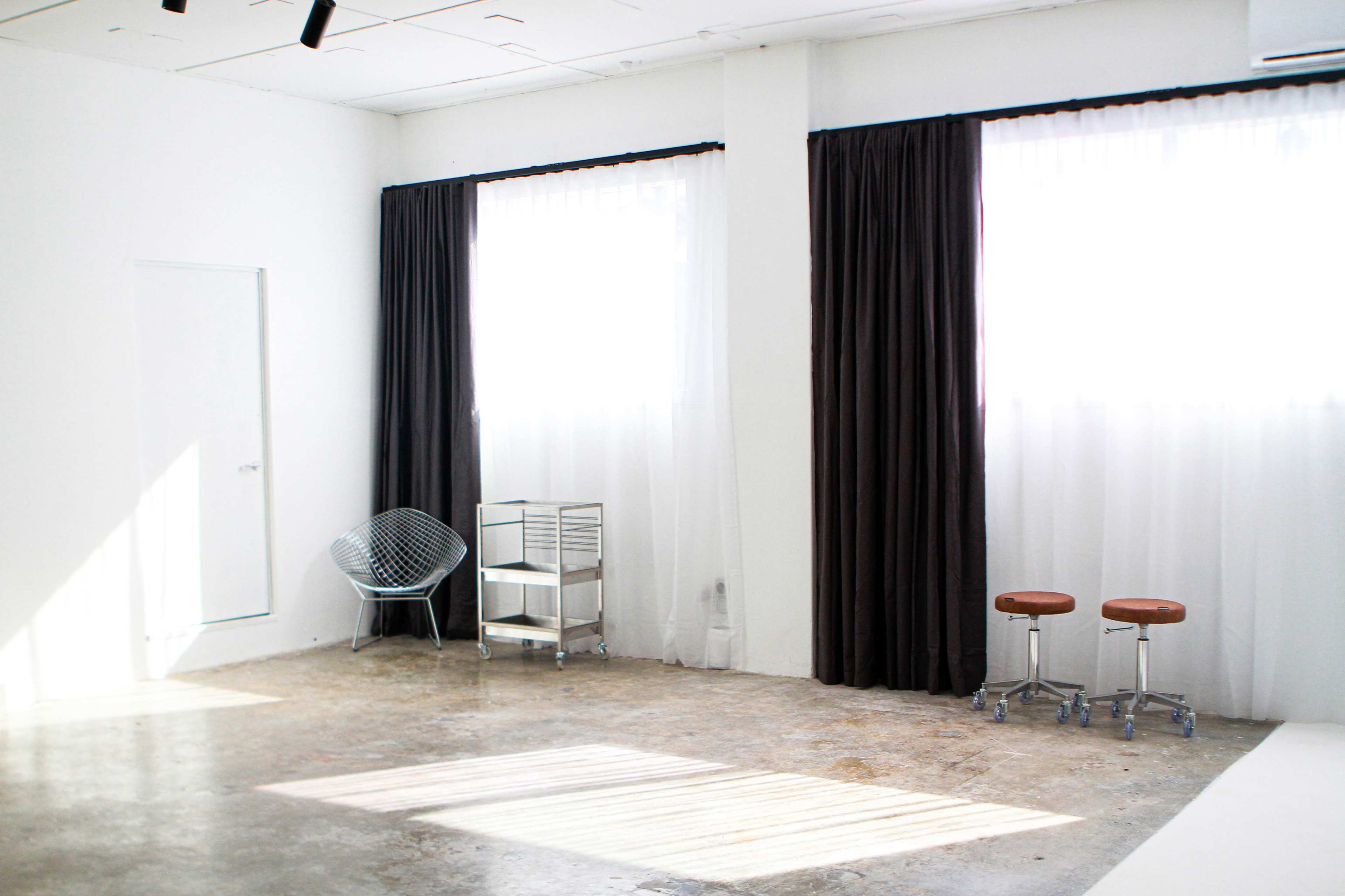 A bright, minimalist room with two large windows, dark curtains, a wire chair, a metal cart, and two stools.