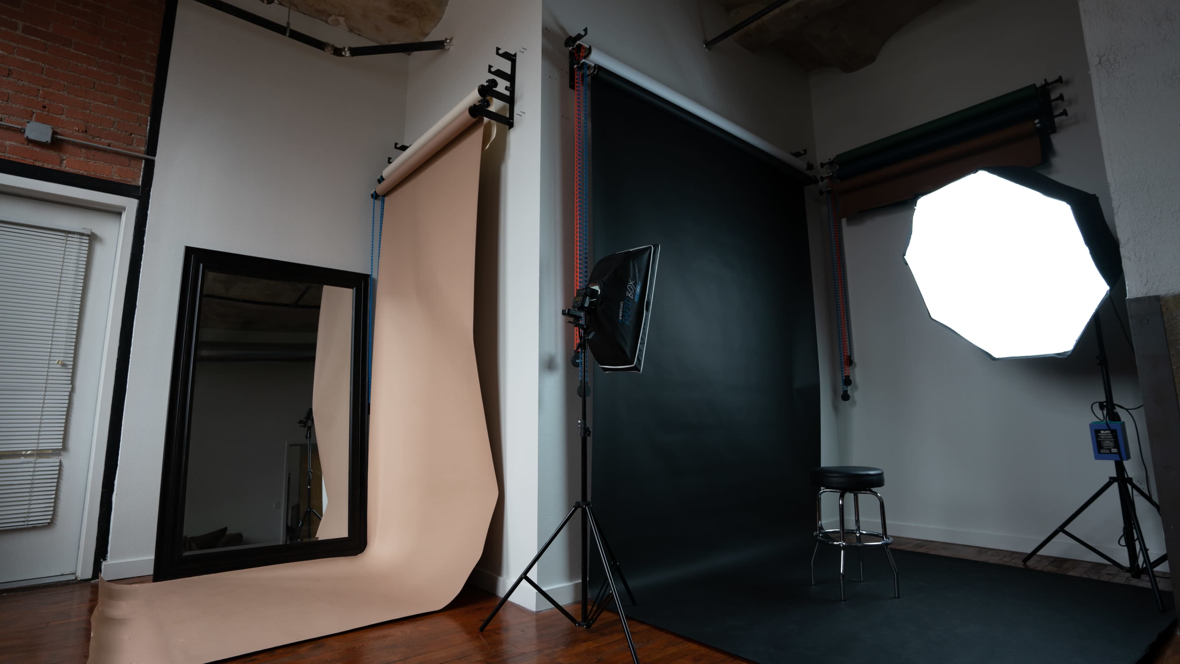 A photography studio with a large mirror, a beige backdrop, a black backdrop, a lighting setup, and a stool.