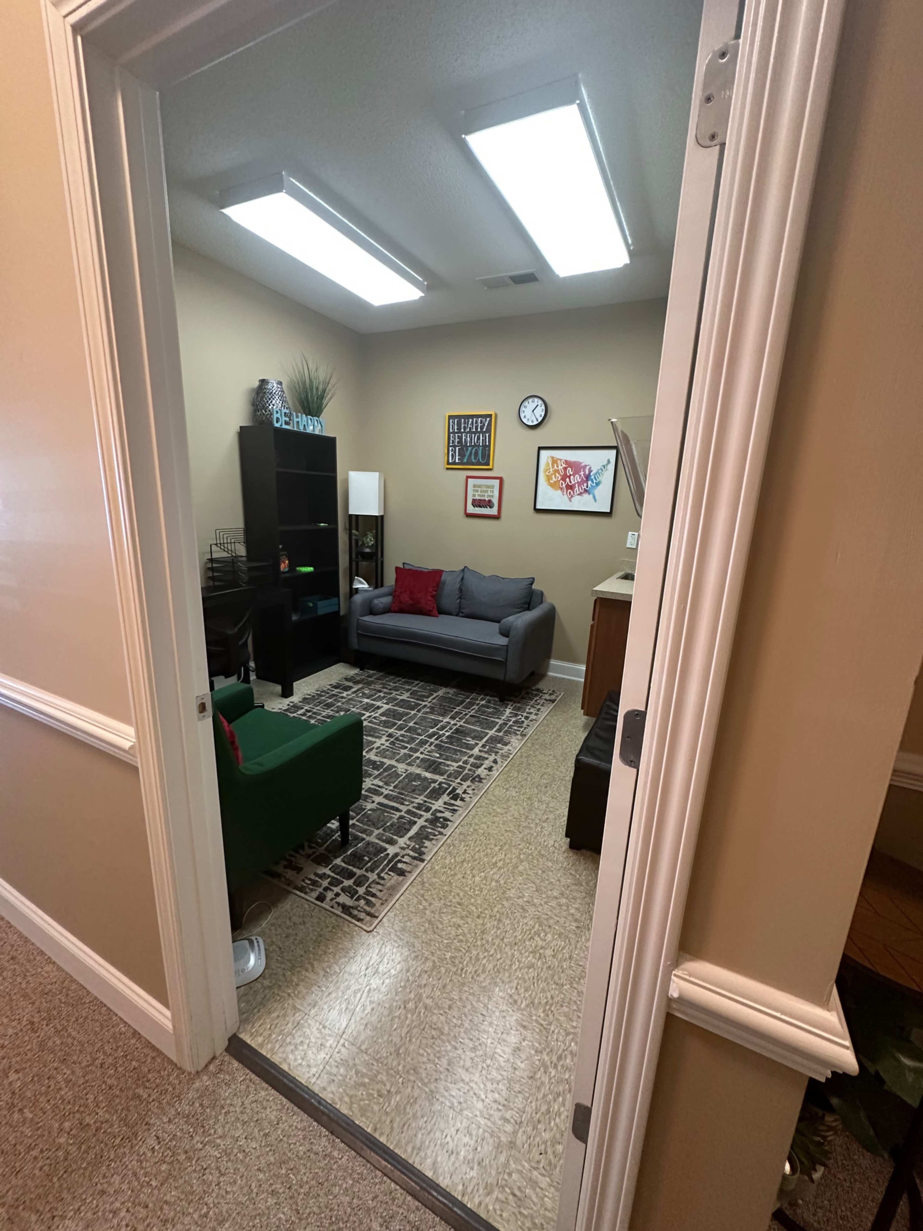 A well-lit room features a gray sofa, a green chair, a bookshelf, and colorful wall art, as seen from an open door.