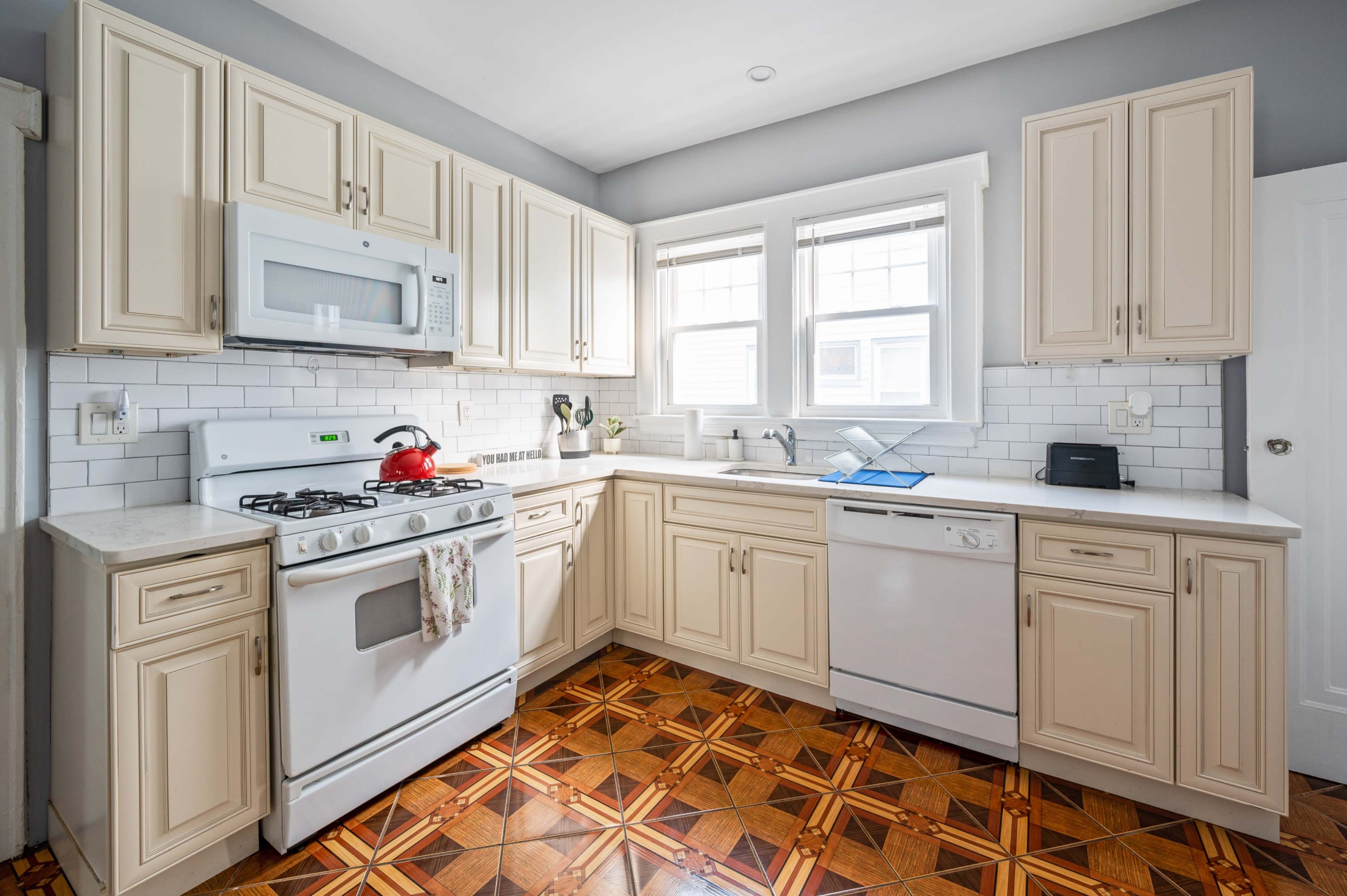 The kitchen features cream-colored cabinets, a gas stove, a microwave, and a dishwasher, with a tiled floor pattern and ample natural light from the windows.
