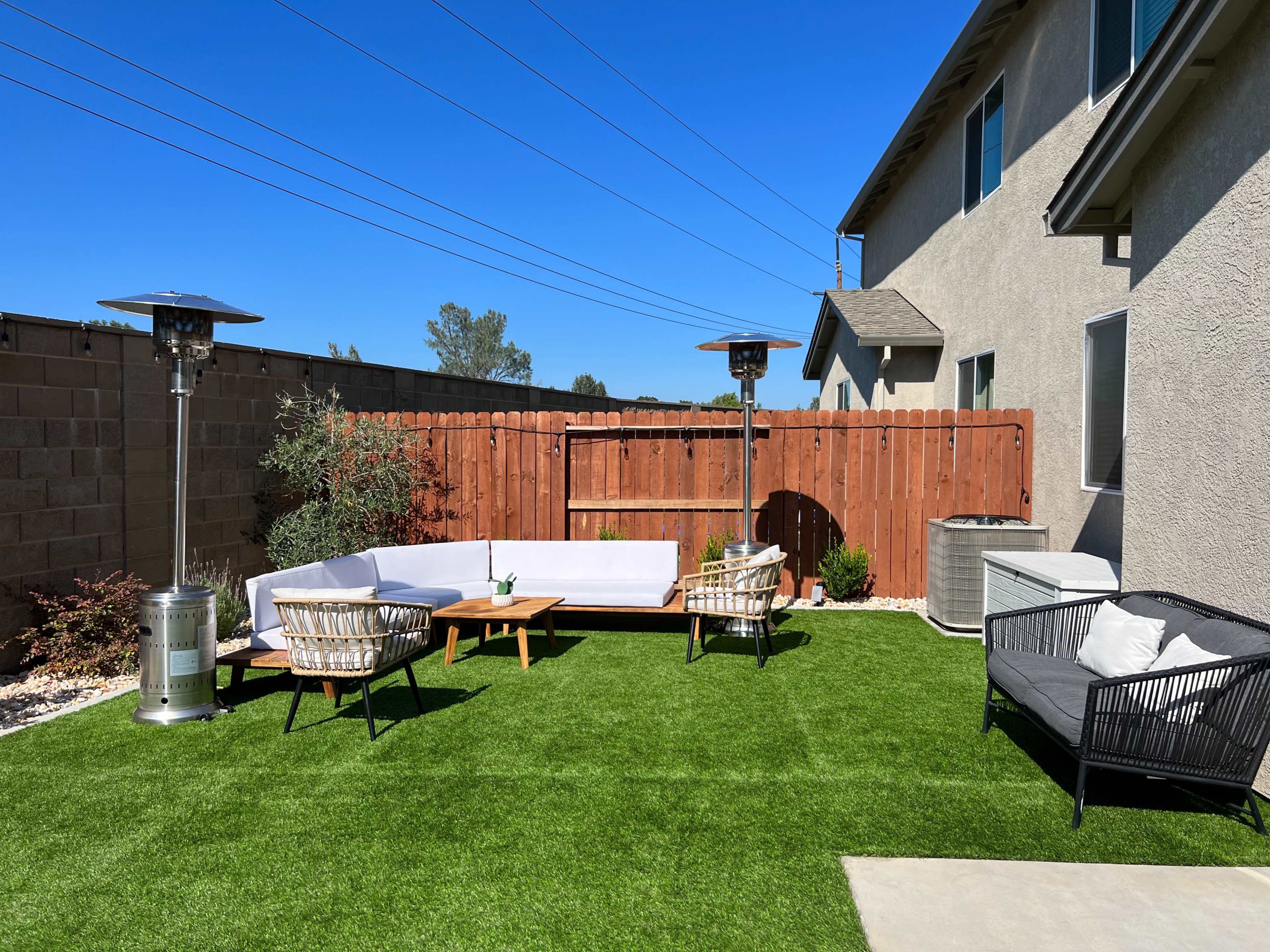 A backyard area with artificial grass, featuring outdoor furniture including a sectional sofa, a coffee table, and two chairs, with propane patio heaters positioned nearby.