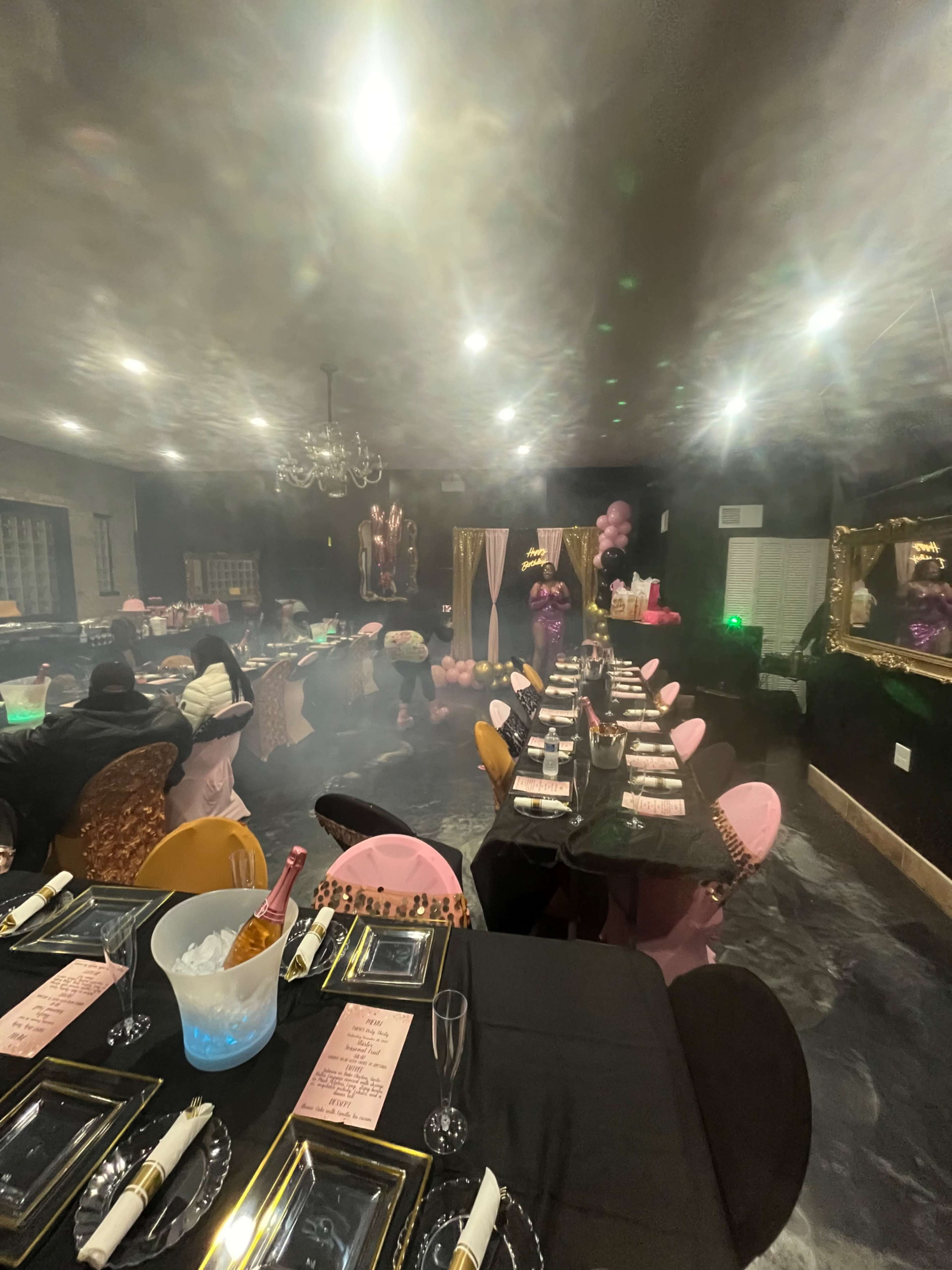 A dimly lit dining room set up for a party, featuring elegantly arranged tables with black tablecloths, colorful chairs, and decorations including balloons and soft lighting.