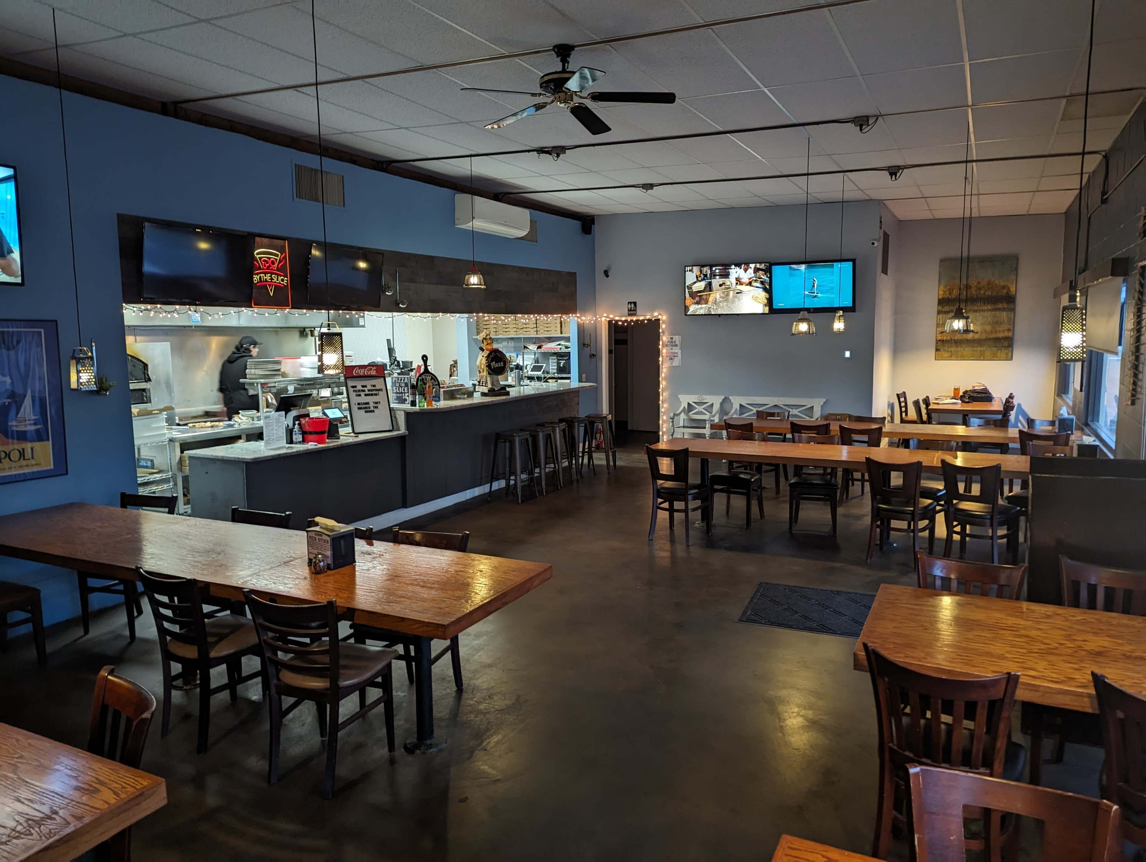 The interior of a casual restaurant with several wooden tables and chairs, an open kitchen in the background, and multiple televisions mounted on the walls.