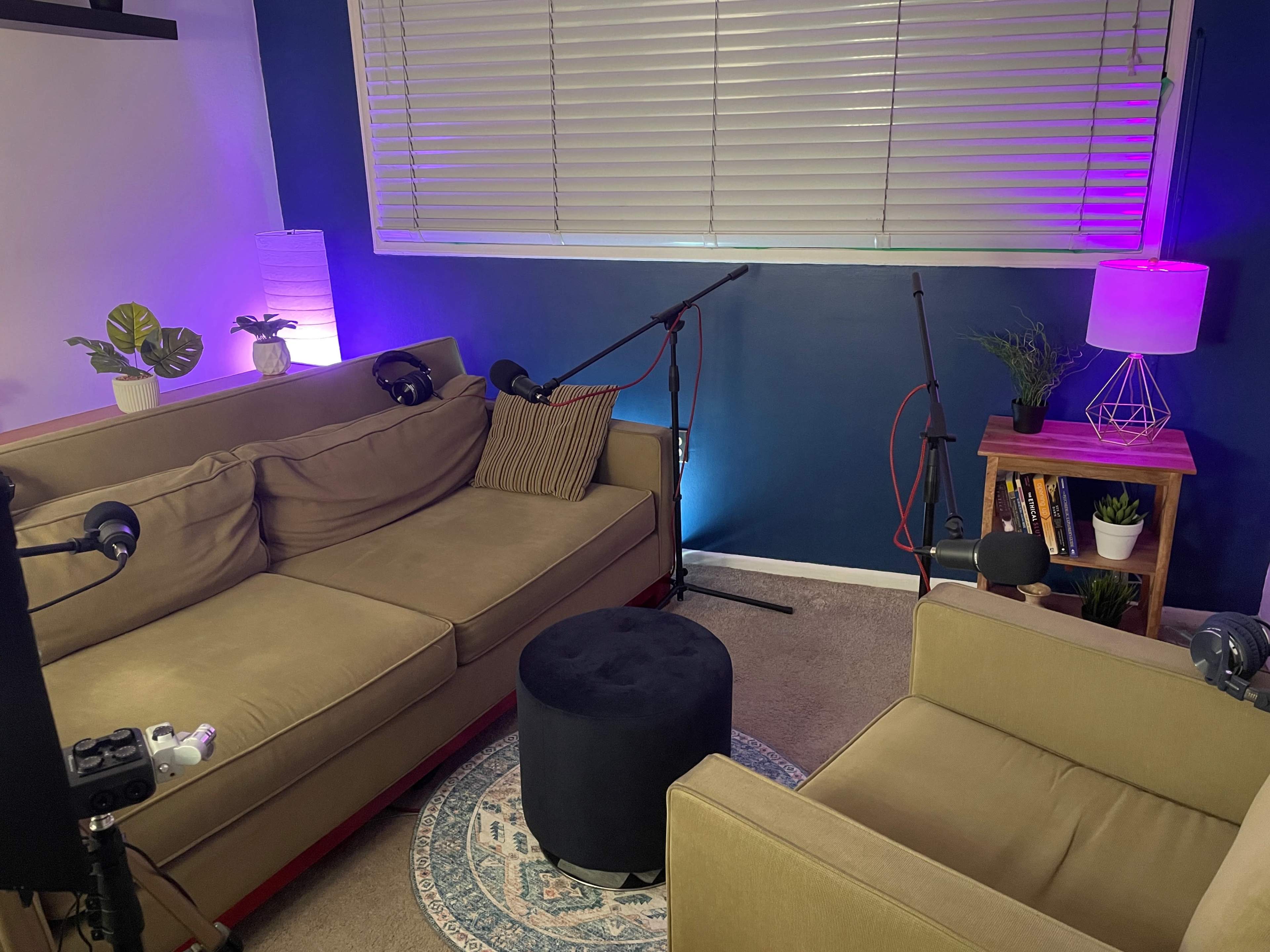 A cozy podcast recording setup features two sofas, a round black ottoman, and microphones positioned on stands, illuminated by ambient lighting from lamps.