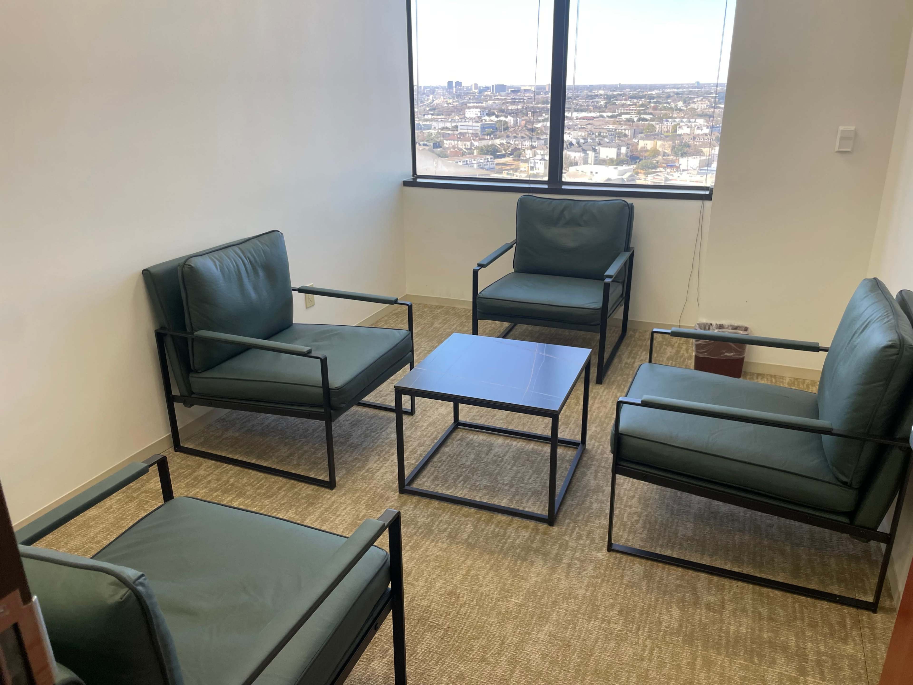 The image shows a small meeting room with four green chairs and a square table, overlooking a city skyline through a large window.