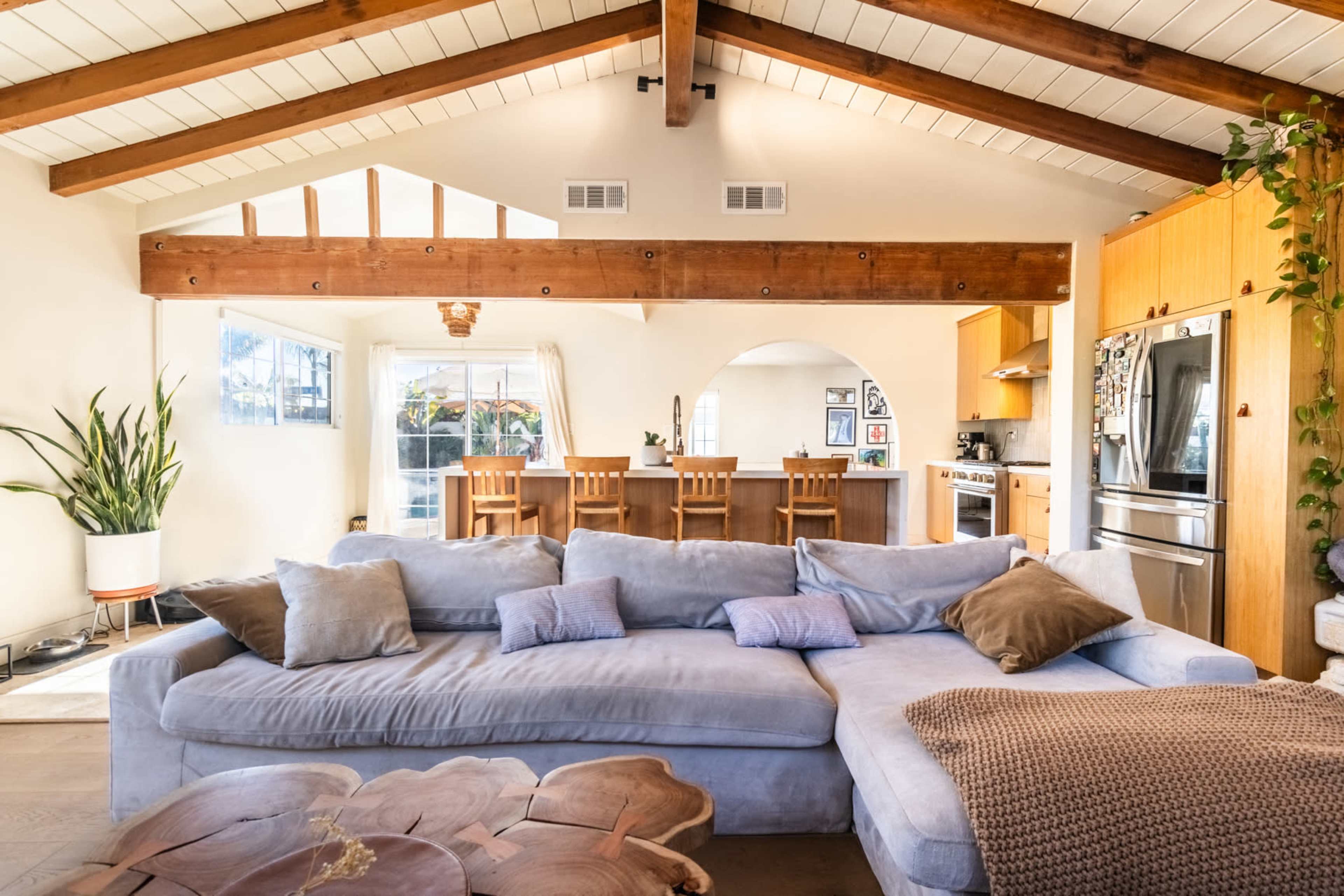 A spacious living room with a large sectional sofa, wooden beams on the ceiling, and a well-lit kitchen area in the background.