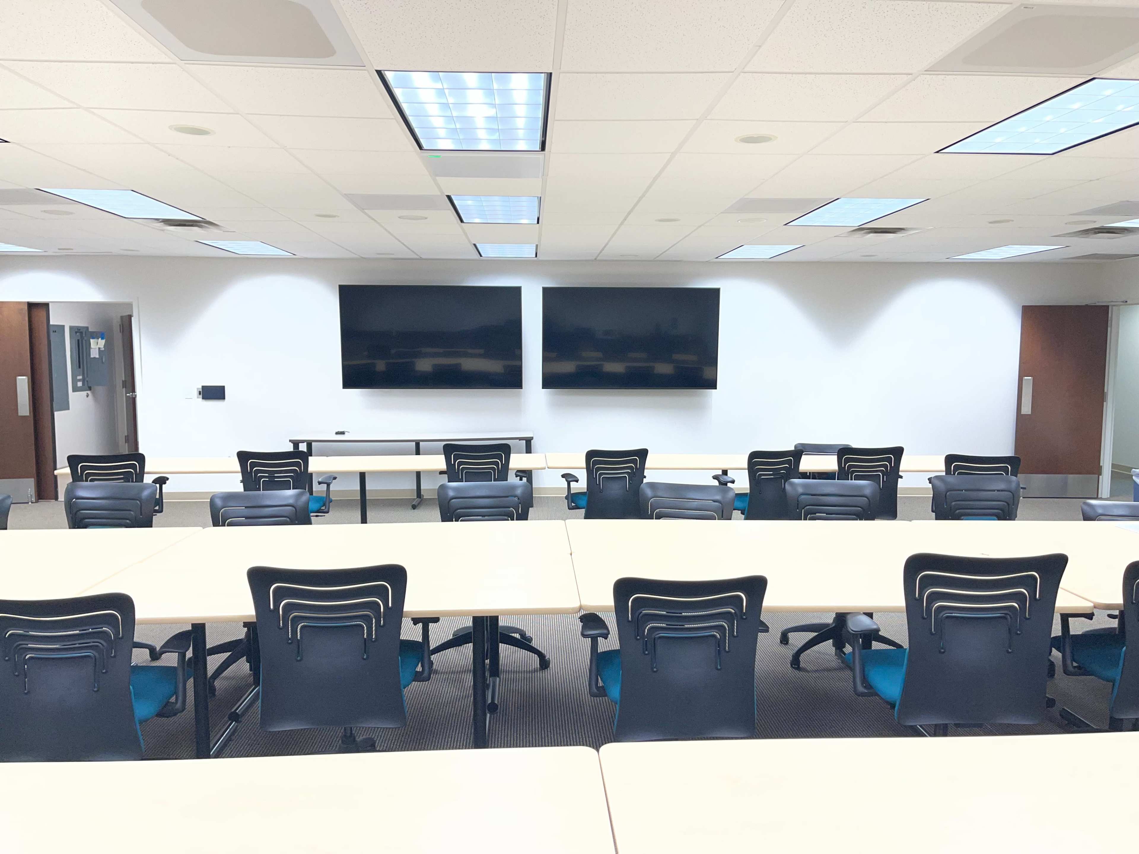 The image shows a modern classroom or meeting room with multiple rows of tables and chairs, two large screens mounted on the wall, and bright ceiling lights.