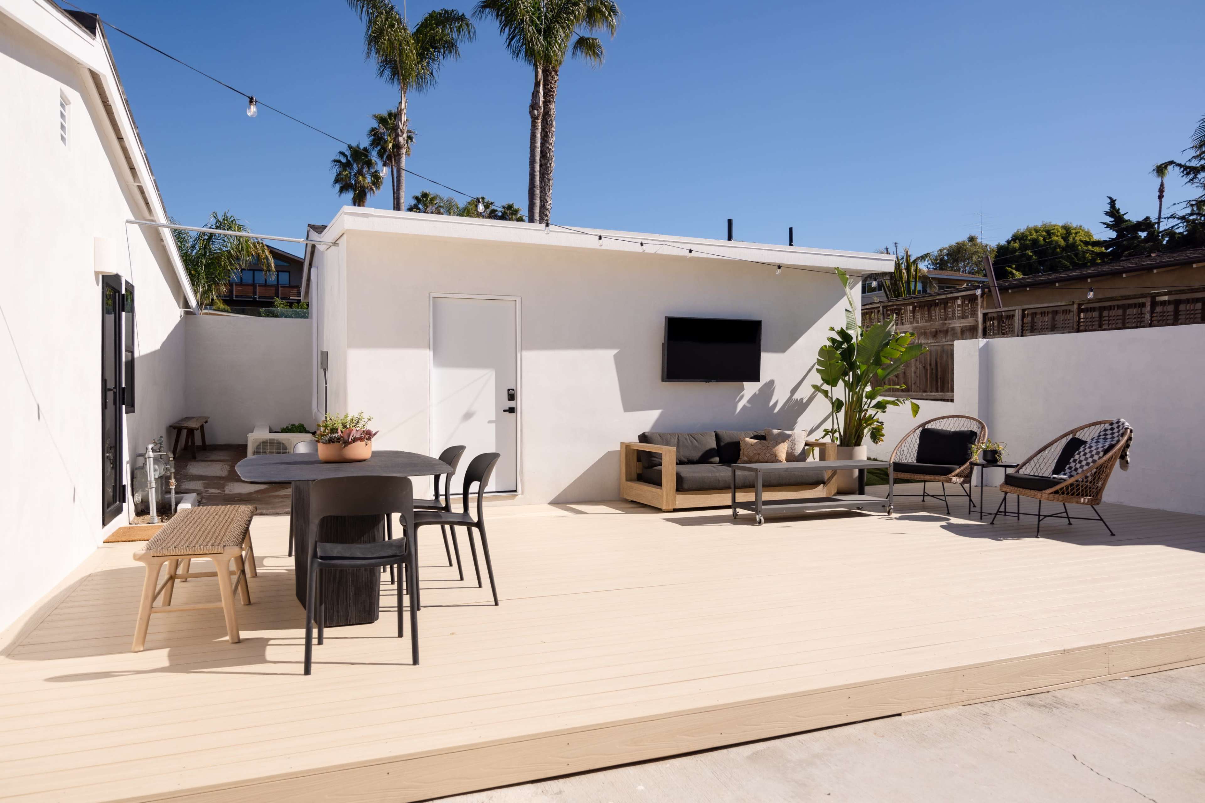 The image shows a modern outdoor patio with a wooden deck, featuring a dining table, lounge area, and potted plants against a backdrop of palm trees.