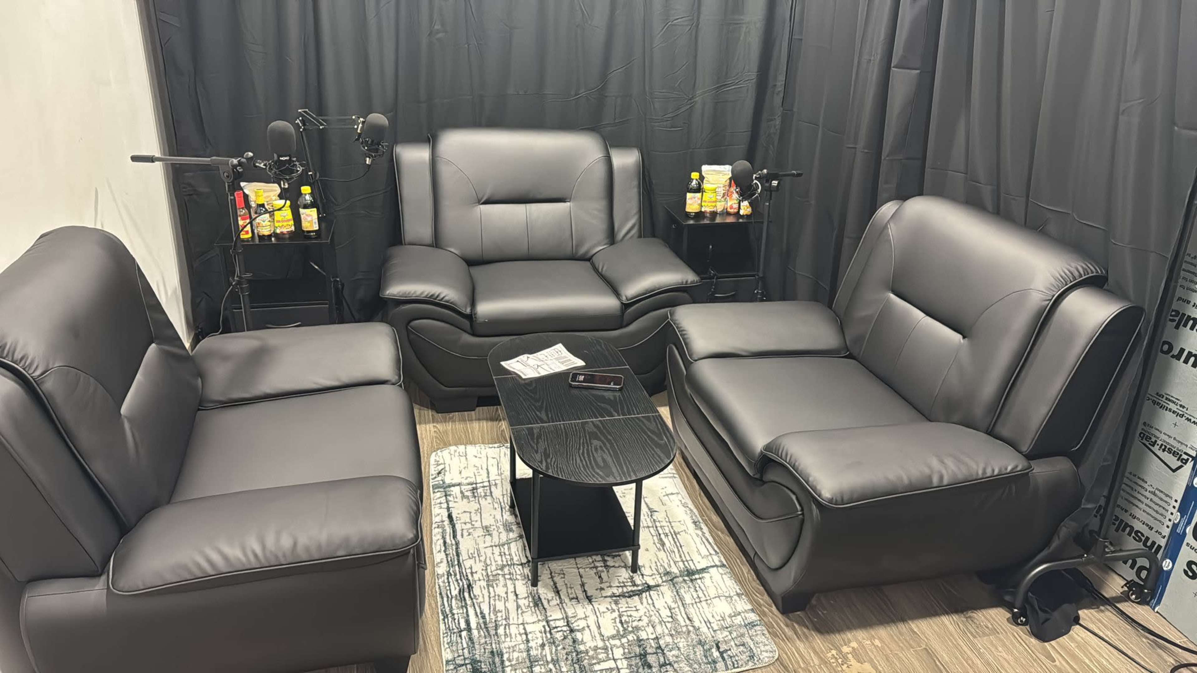 The image shows a small seating area with black leather sofas arranged around a central black coffee table, surrounded by dark curtains and equipped with two microphones on stands.