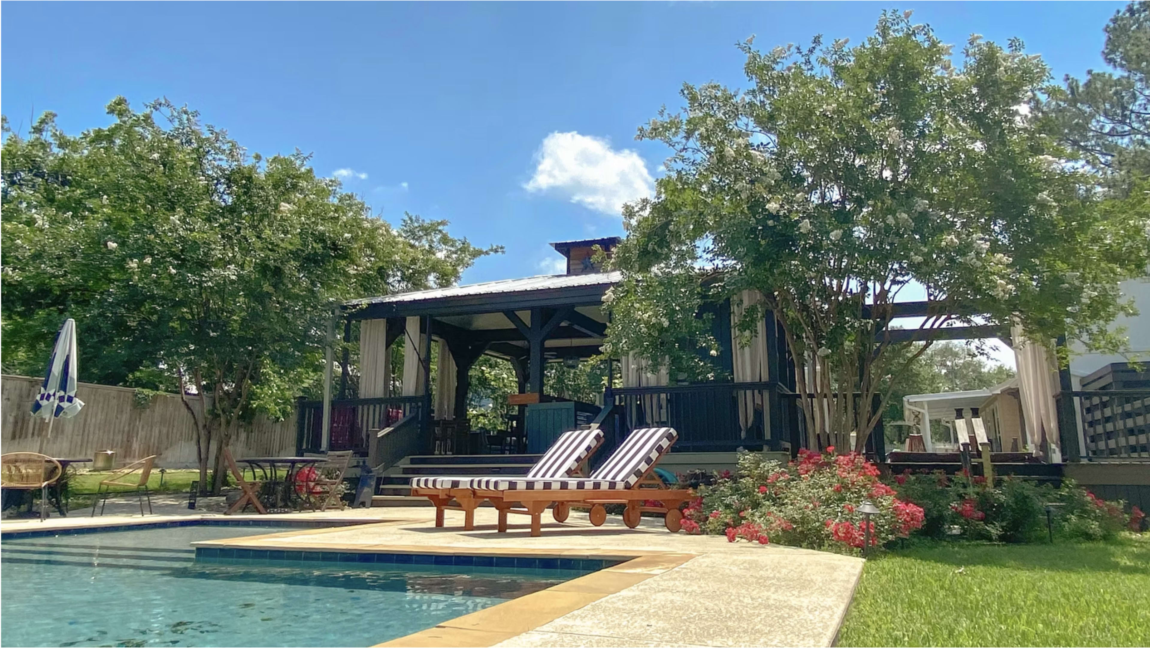 The image shows a swimming pool with lounge chairs and a covered patio surrounded by trees and blooming flowers.