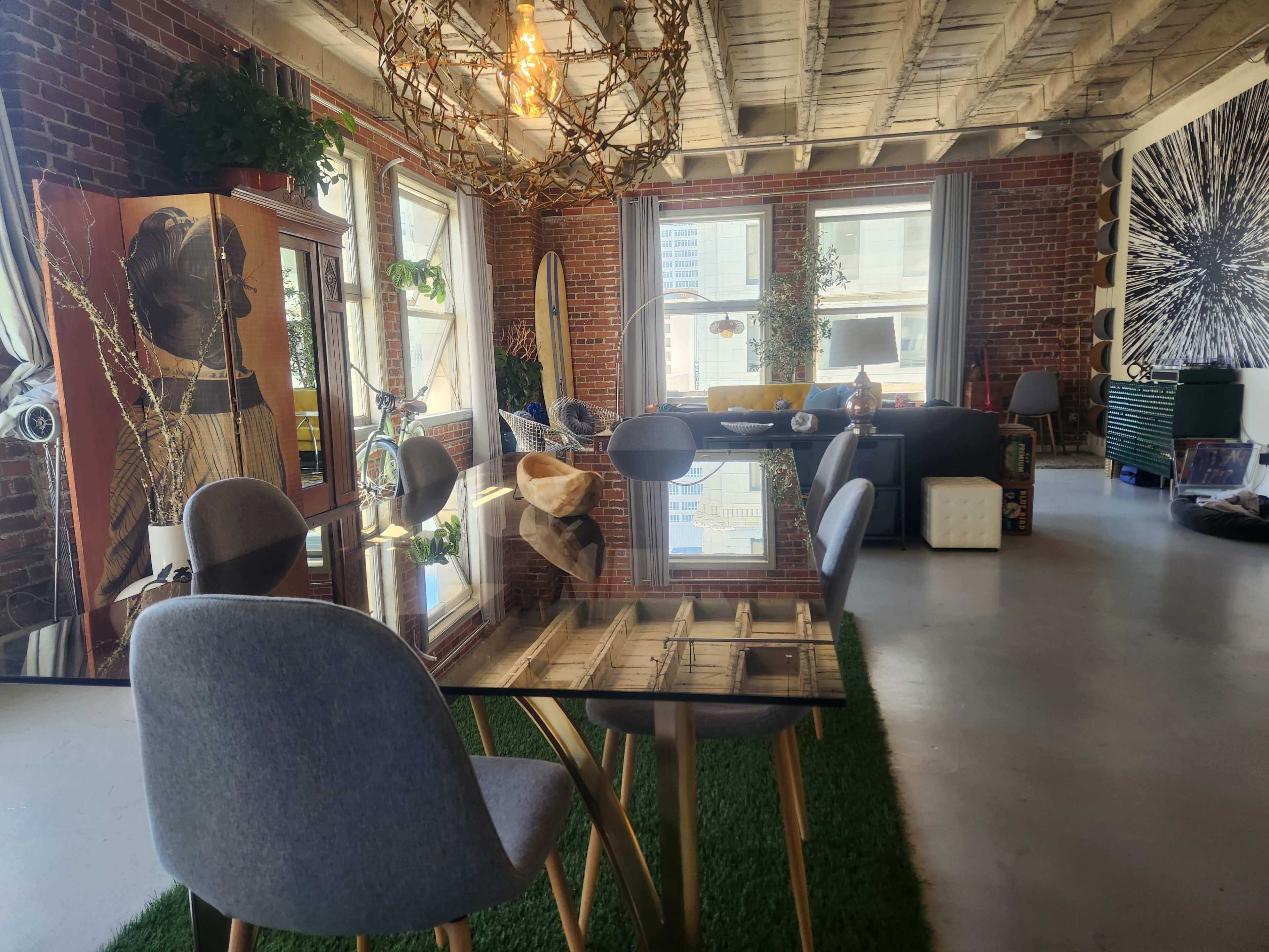 The image depicts a spacious, modern living area with brick walls, large windows, a glass dining table surrounded by gray chairs, and various decorative elements including plants and artwork.