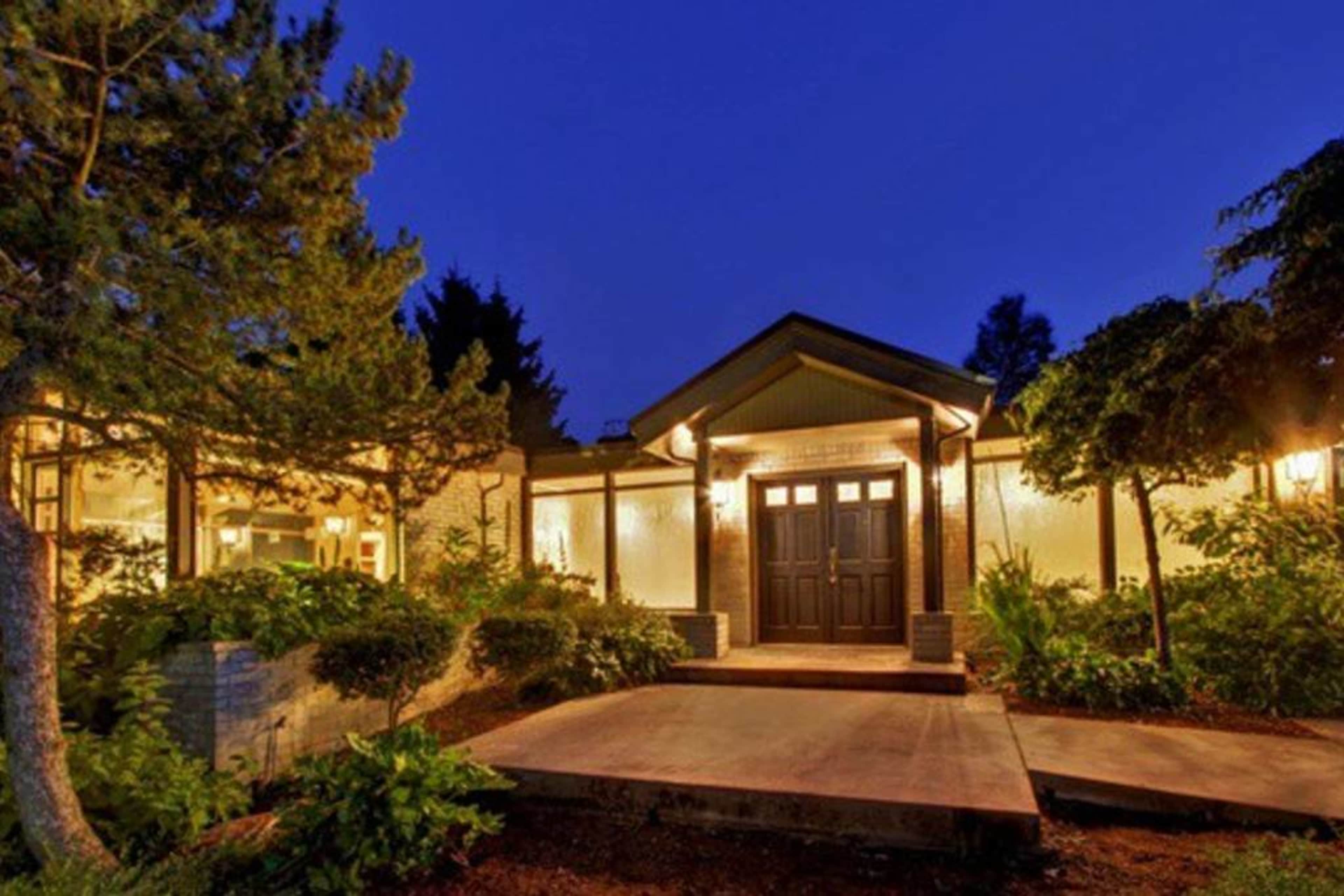 A modern house with a dark front door and warm lighting is surrounded by greenery at dusk.