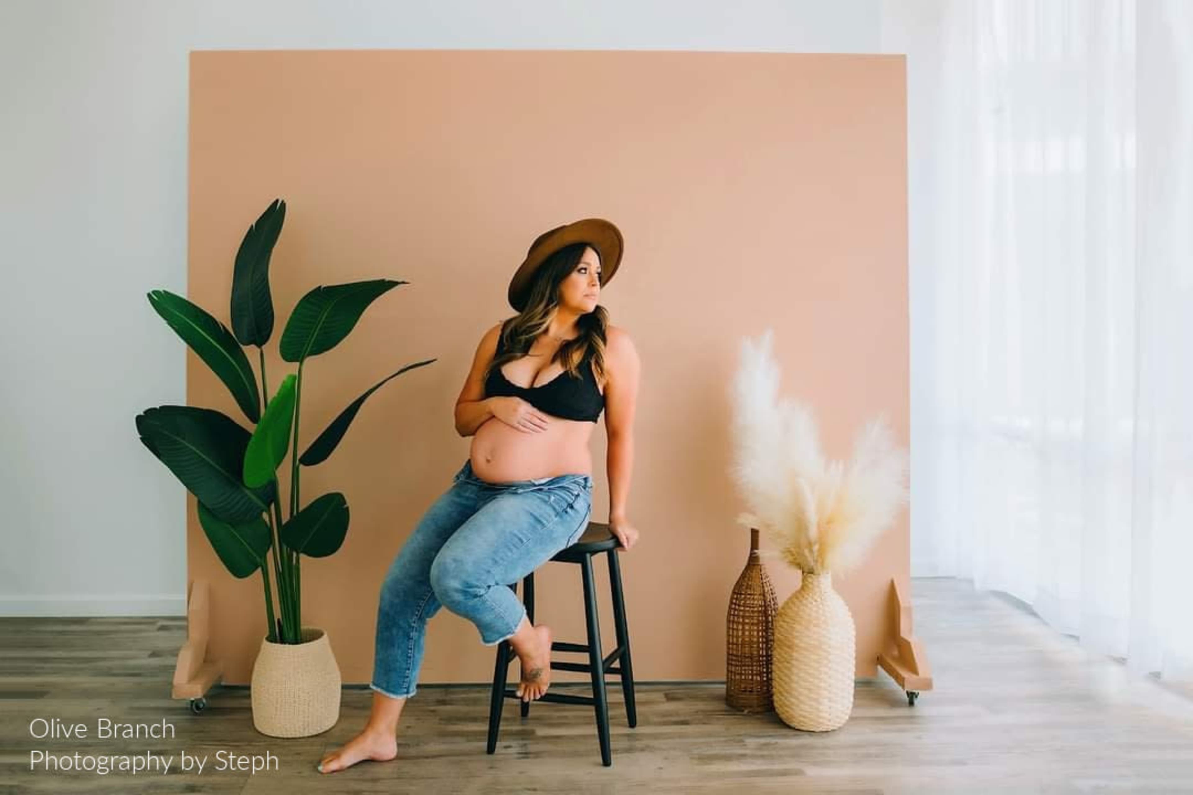 A pregnant woman in a black top and jeans sits on a stool beside a potted plant and decorative vases against a peach-colored backdrop.