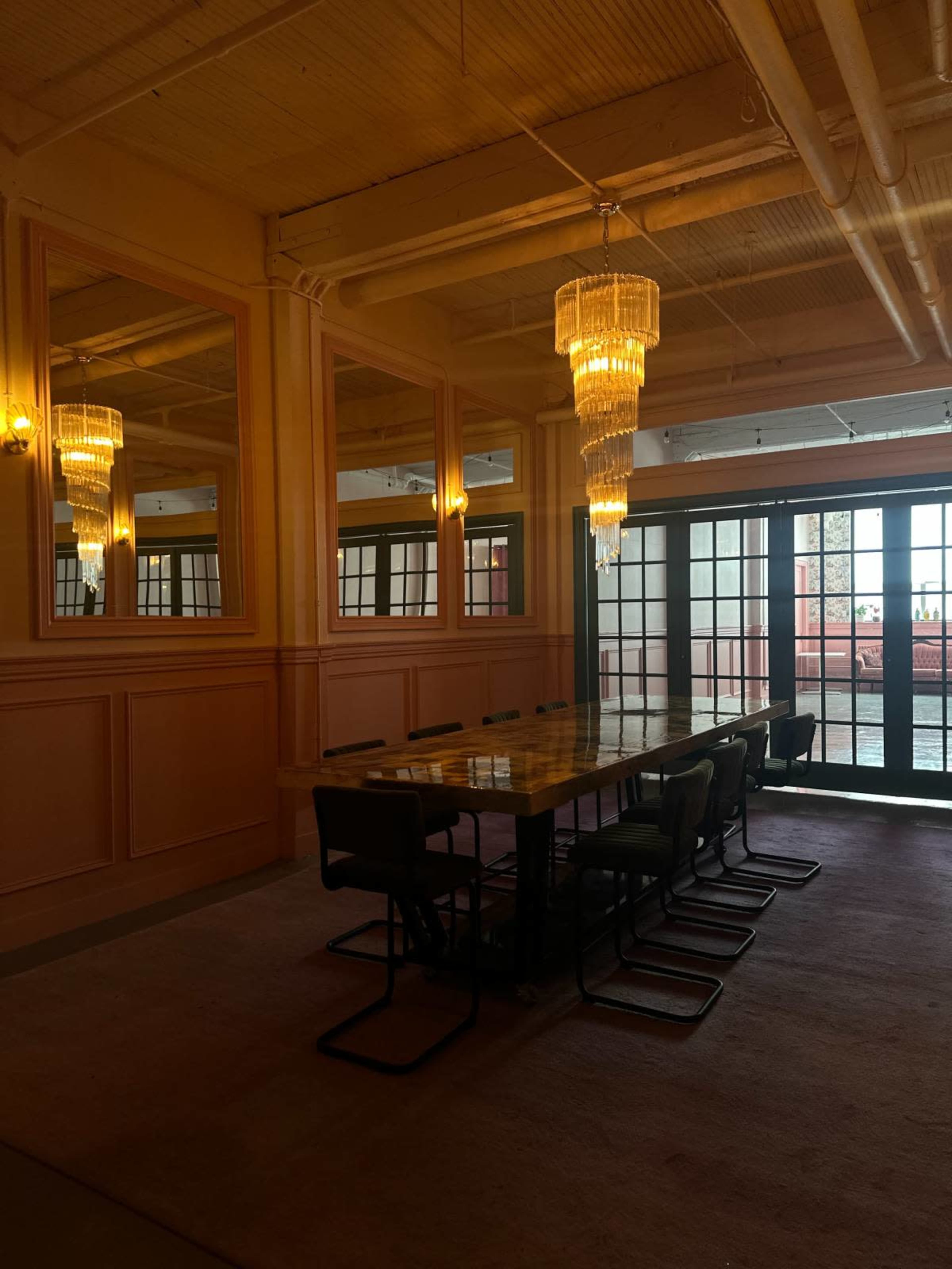 A dining room features a long table surrounded by chairs, illuminated by two ornate chandeliers and flanked by large mirrors on the walls.