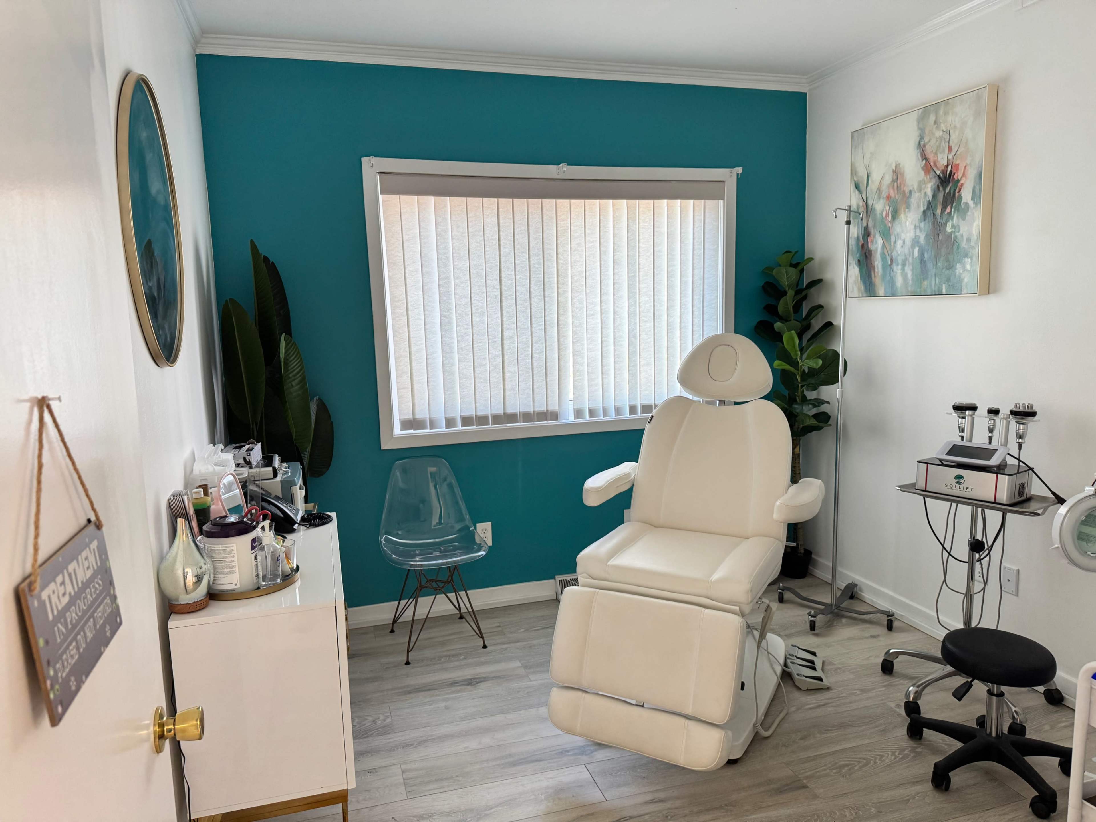 The image shows a well-lit treatment room featuring a white reclining chair, a clear acrylic chair, and various aesthetic equipment along with teal walls and a window covered by sheer blinds.