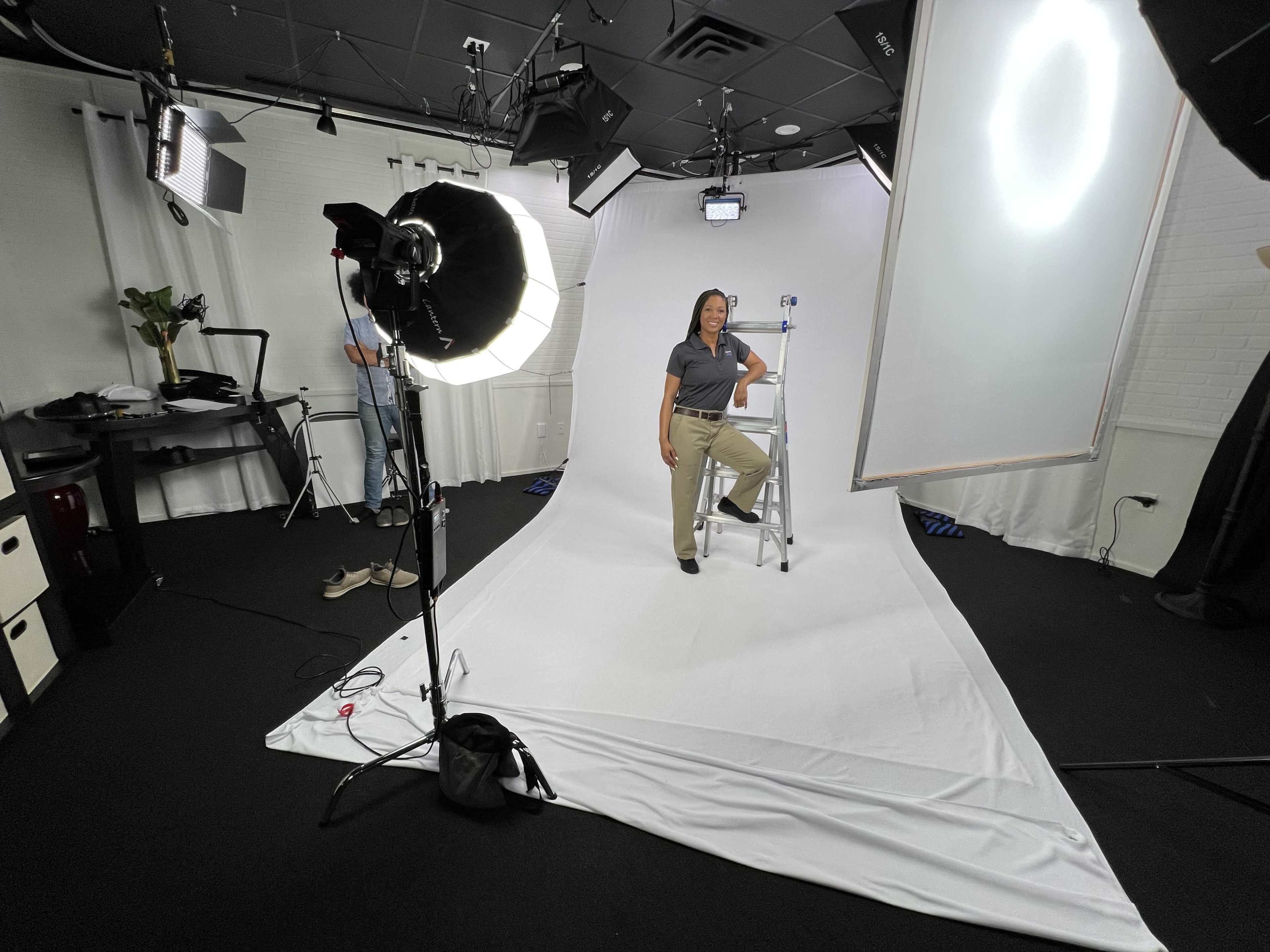 A woman sits on a ladder in a studio setup with lighting equipment and a backdrop.