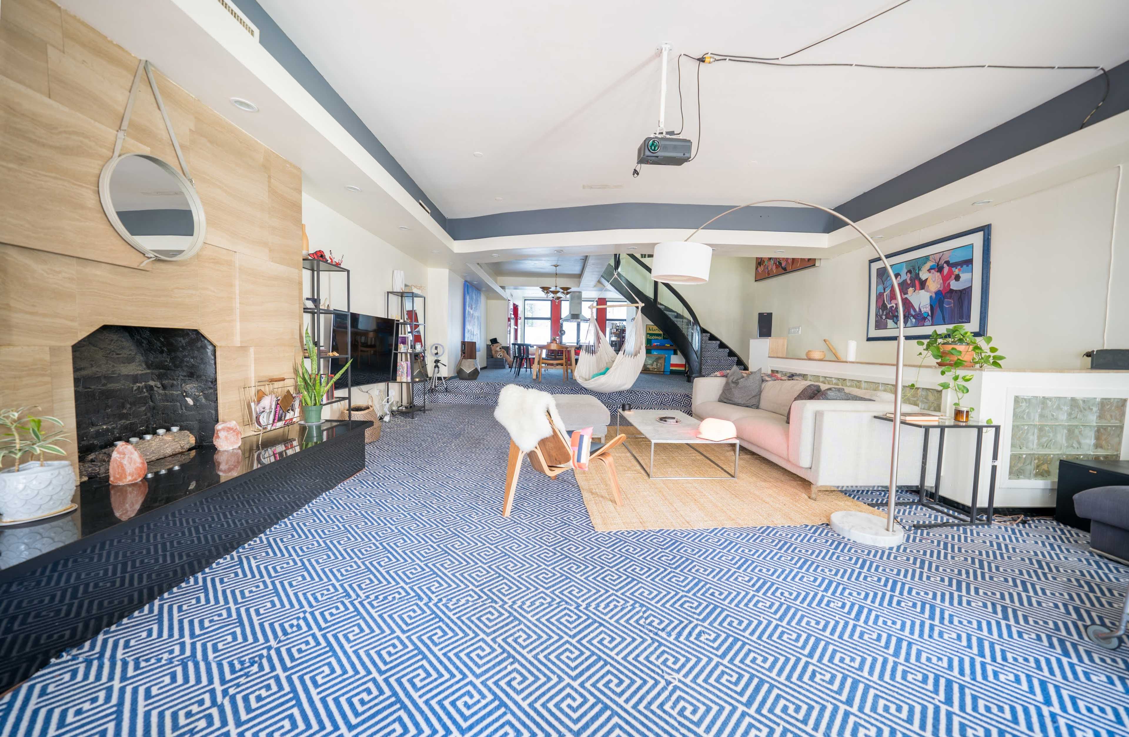 The image shows a spacious living room with a blue and white patterned carpet, a modern fireplace, a large sectional sofa, and a hanging hammock.