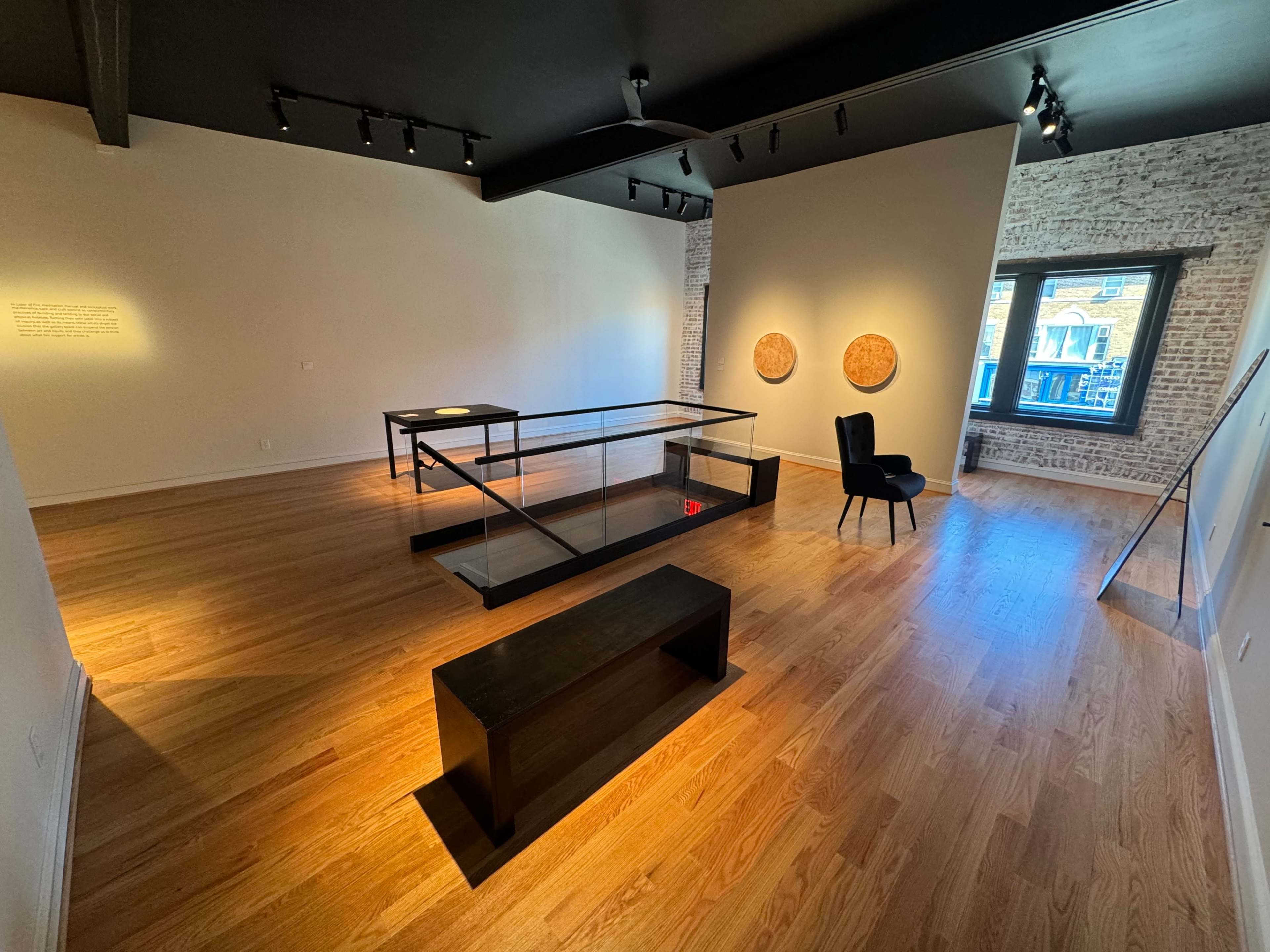 The image shows a minimalist art gallery with wooden flooring, a glass display case in the center, a black chair against a wall, and two circular artworks mounted on the opposite wall.