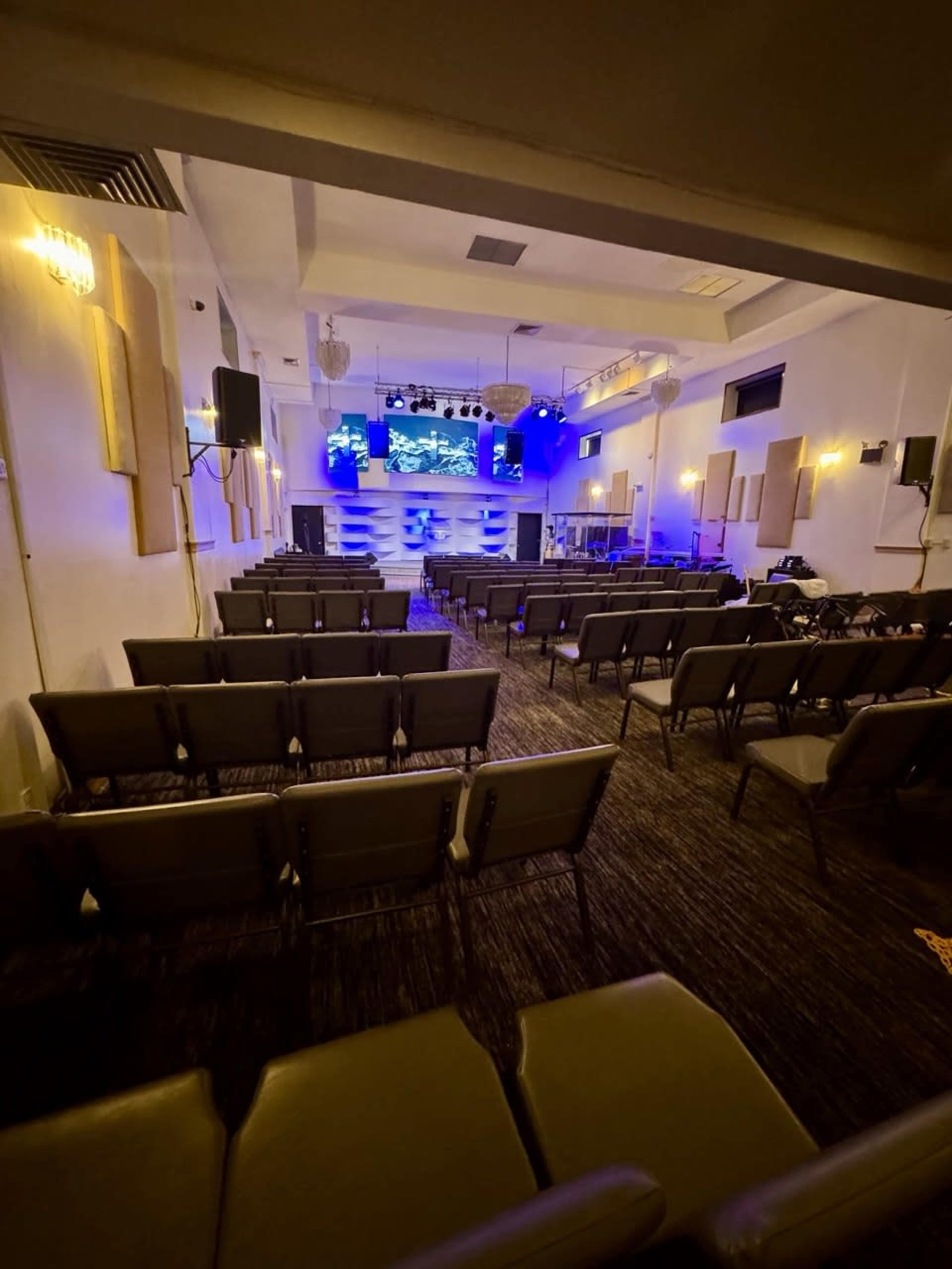 The image shows an interior view of a modern auditorium with rows of empty chairs facing a stage illuminated by blue lighting and featuring a large screen.