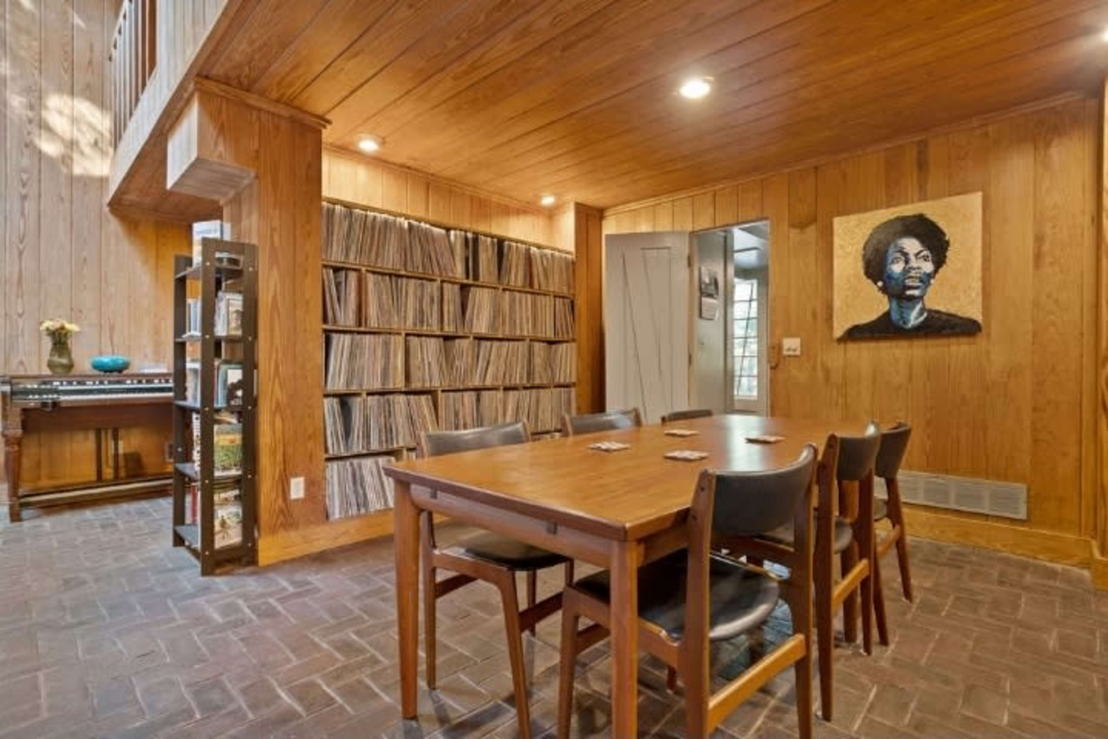 The scene features a wooden dining area with a table and chairs, adjacent to a wall lined with vinyl records and a portrait of a woman.