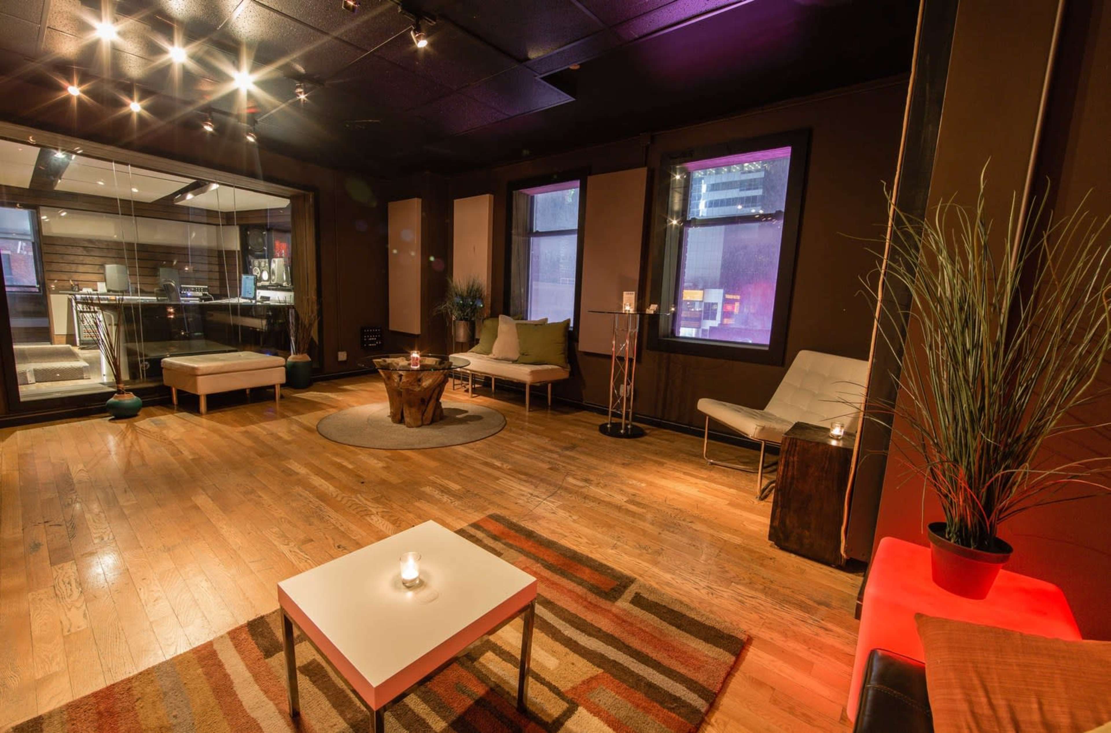The image shows a spacious lounge area with wooden floors, contemporary furniture, and large windows, featuring a central coffee table and a decorative plant.