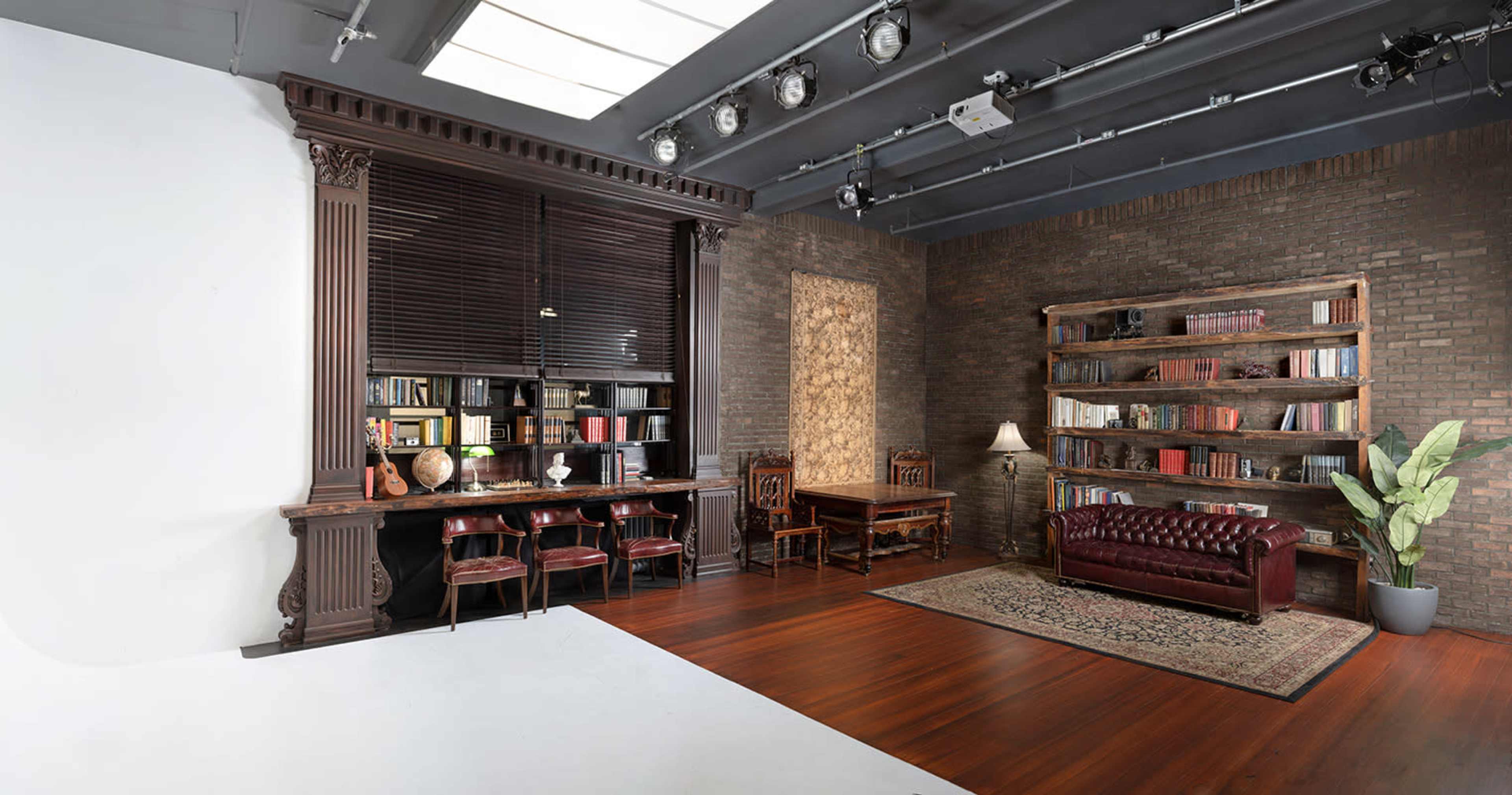 A modern studio space featuring a wooden desk and chairs, a bookshelf filled with books, a leather couch, and a plant, all arranged against a brick wall.
