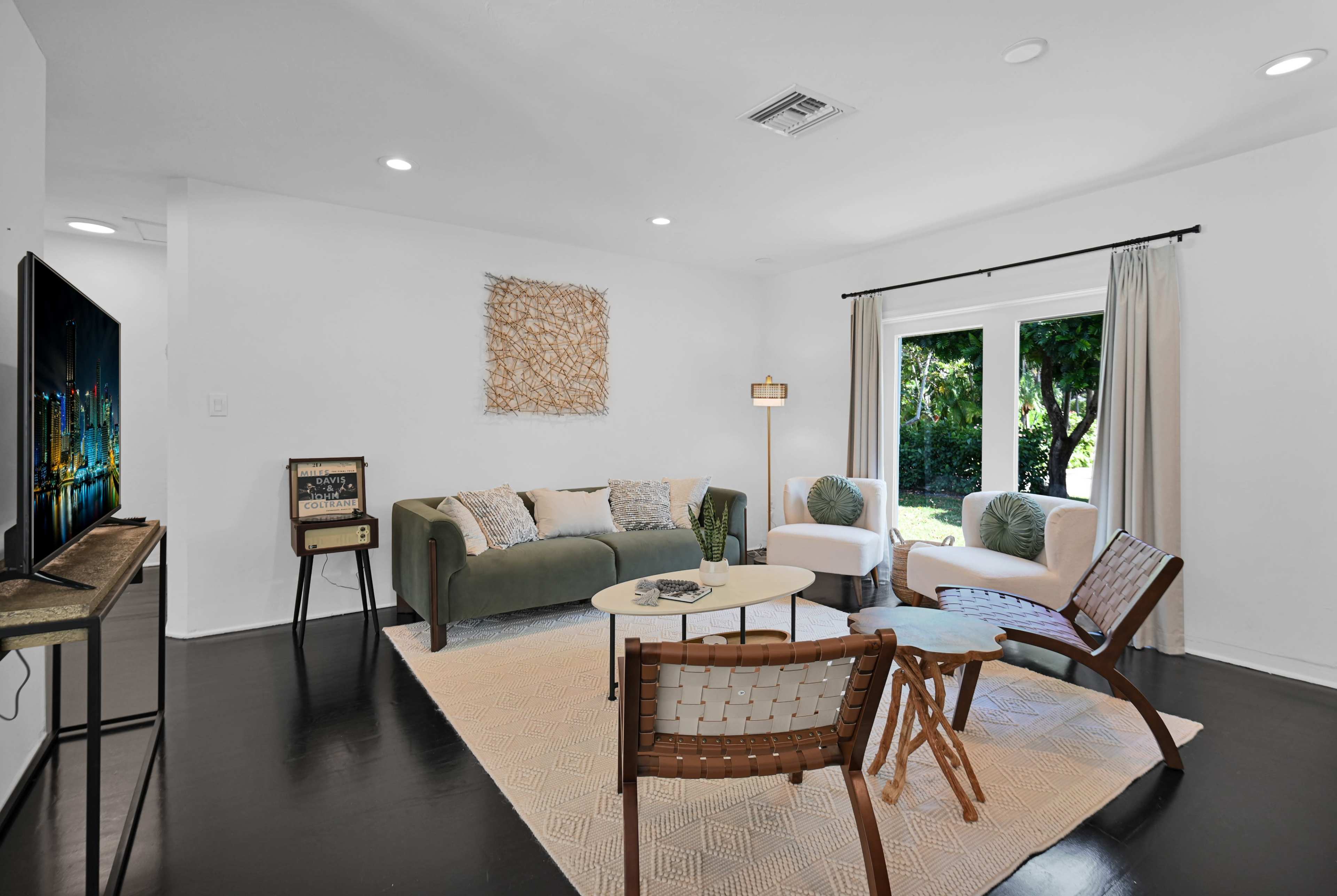 A modern living room with a green sofa, a round coffee table, and various seating options, including wicker chairs, arranged around a large window that opens to a view of greenery.