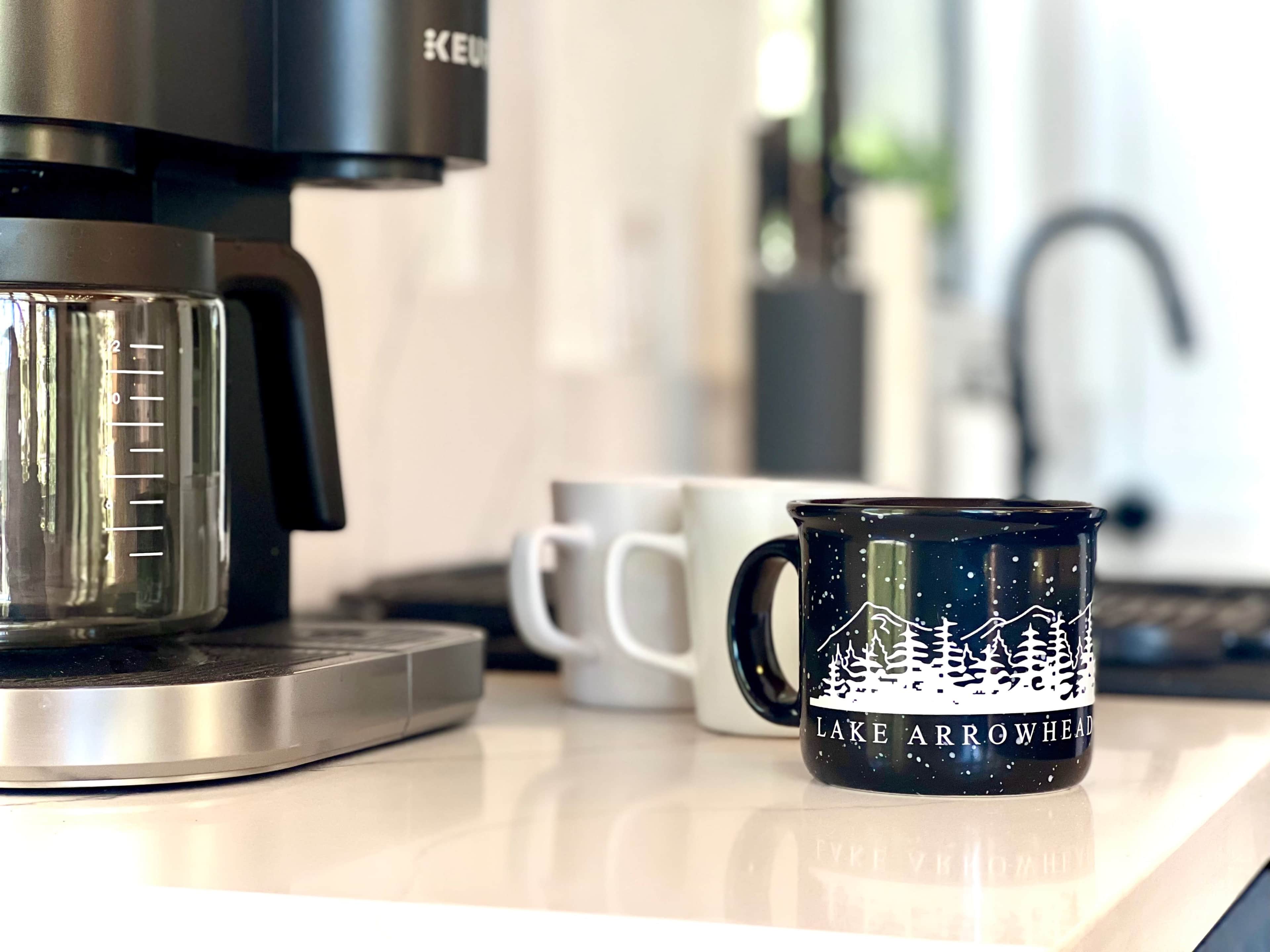 A coffee maker sits on a countertop next to several mugs, including a blue one with a "Lake Arrowhead" design.