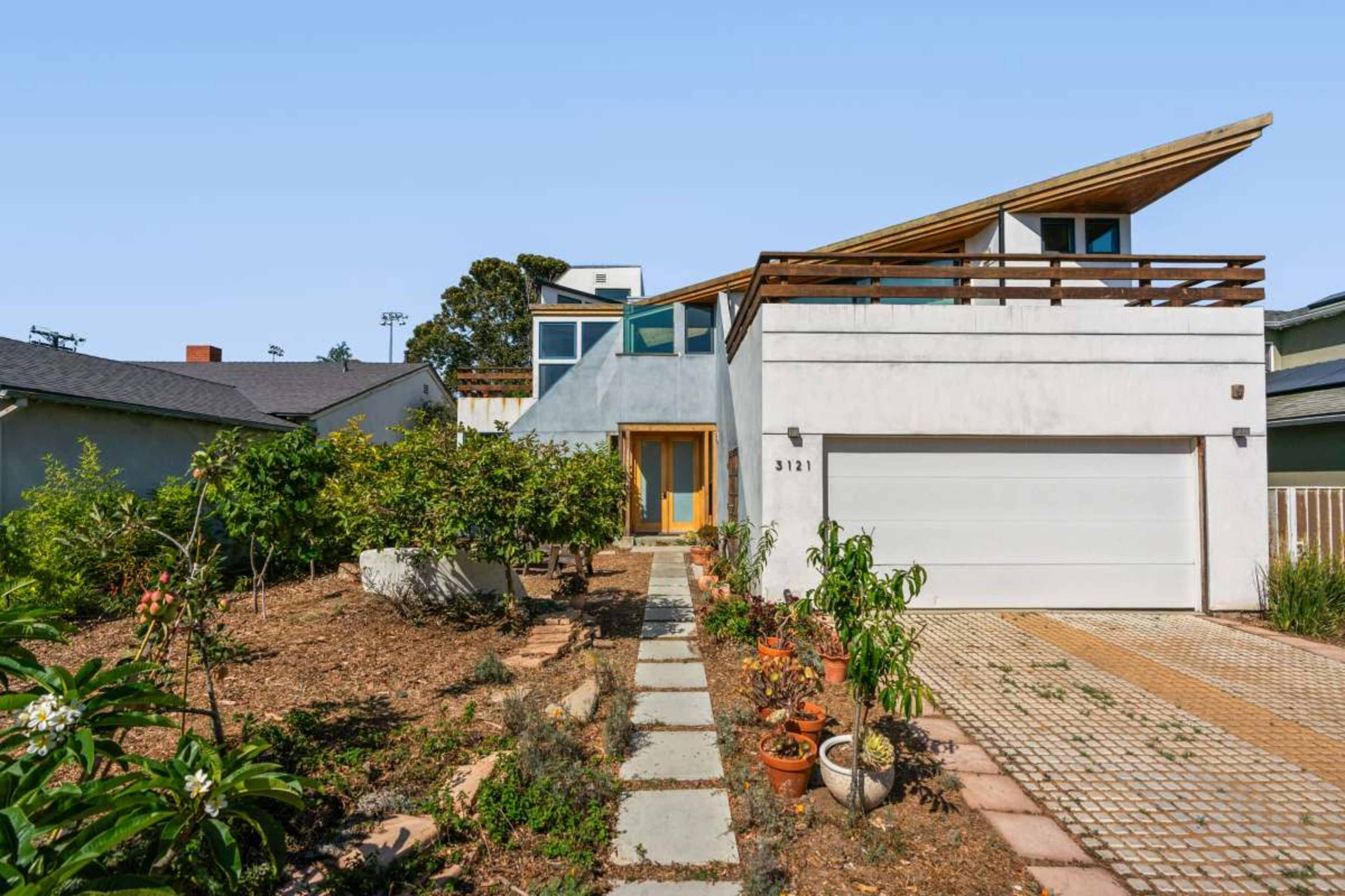 The image shows a modern two-story house with a sloped roof, a garage, and a landscaped front yard featuring potted plants and a stone pathway.