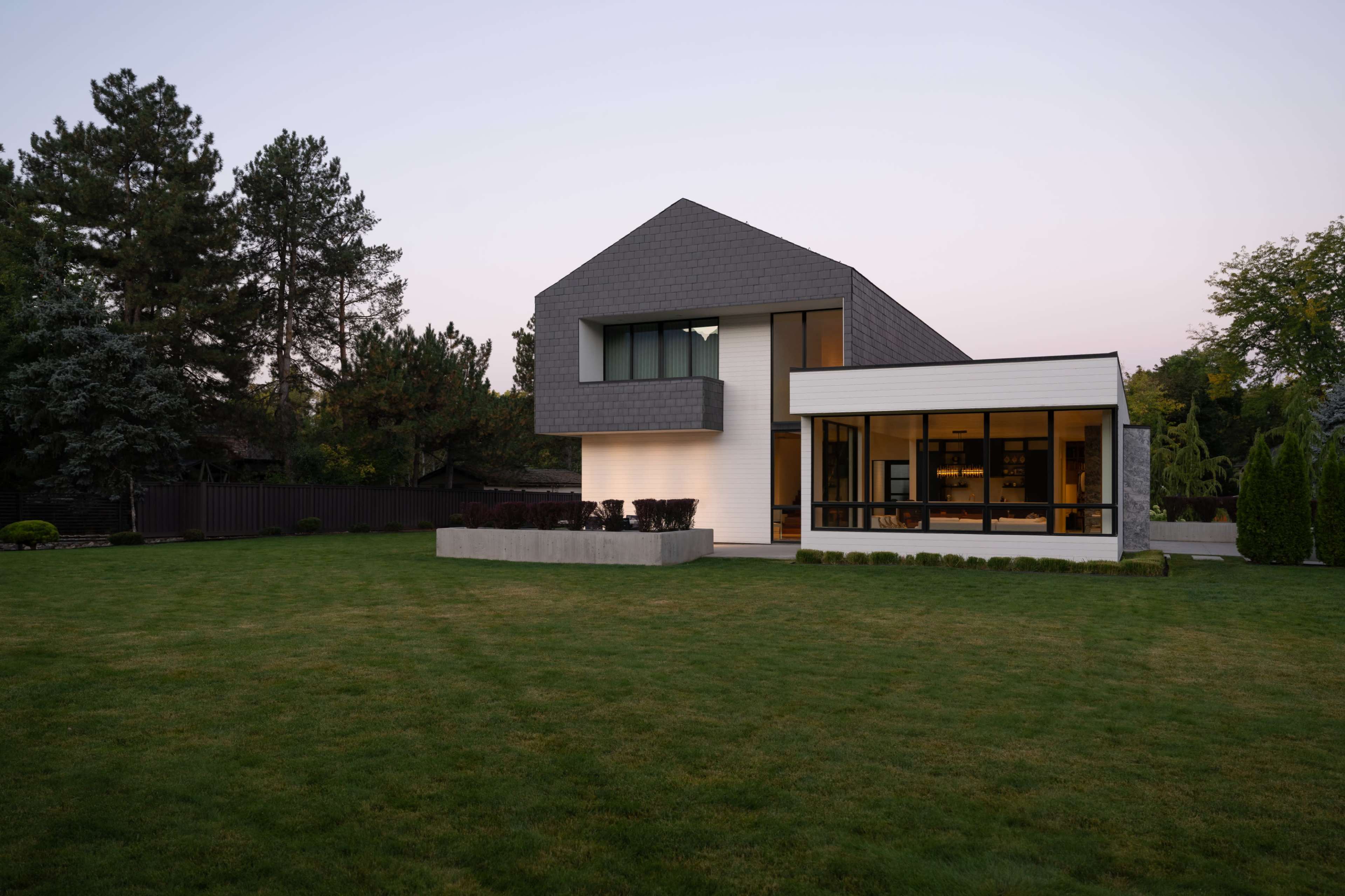 A modern two-story house with a geometric design, featuring a mix of materials, large windows, and a well-maintained lawn is set against a twilight sky.