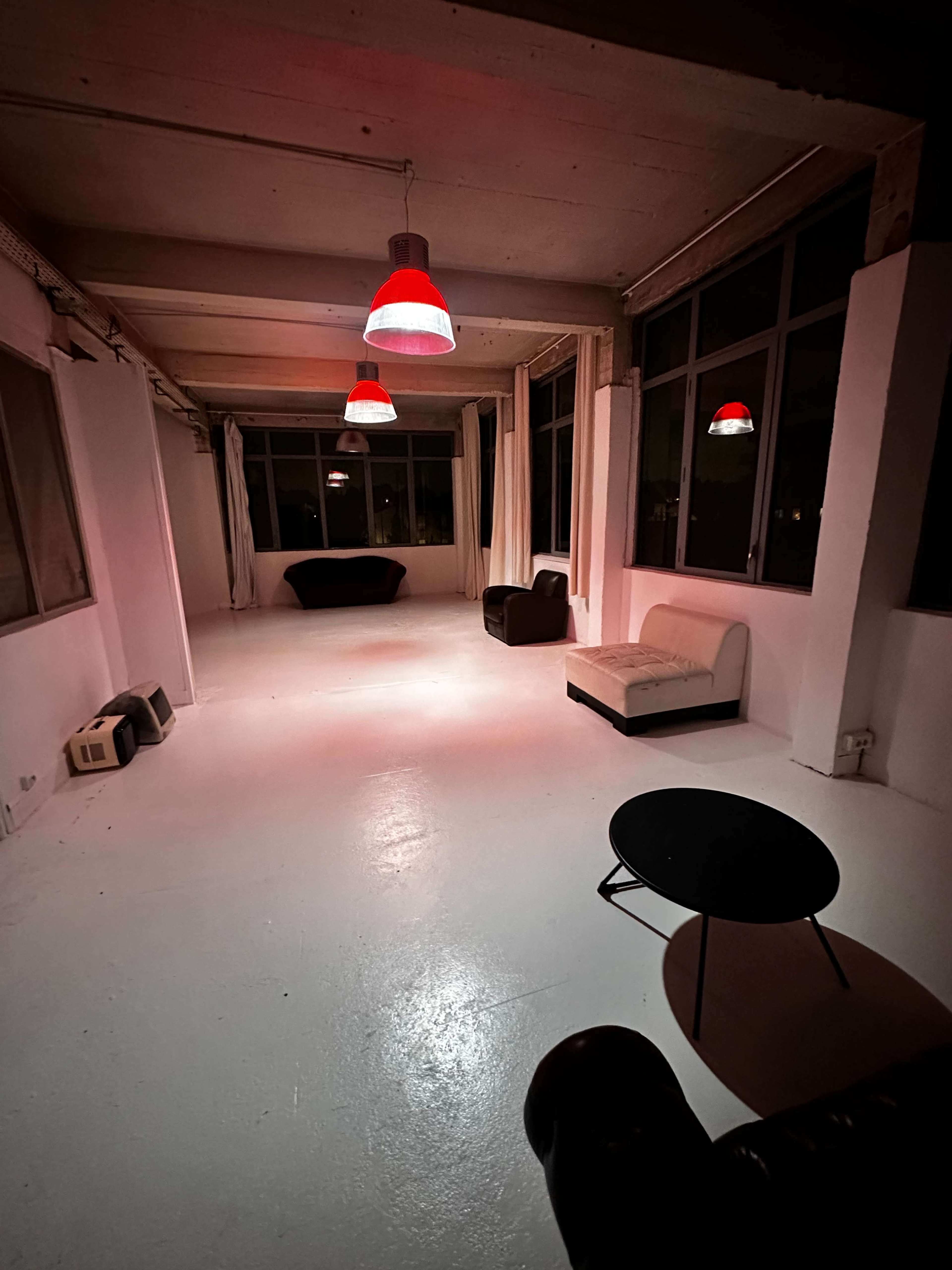 The image shows a spacious room with white floors and large windows, featuring two dark sofas, a small black table, and red pendant lights hanging from the ceiling.