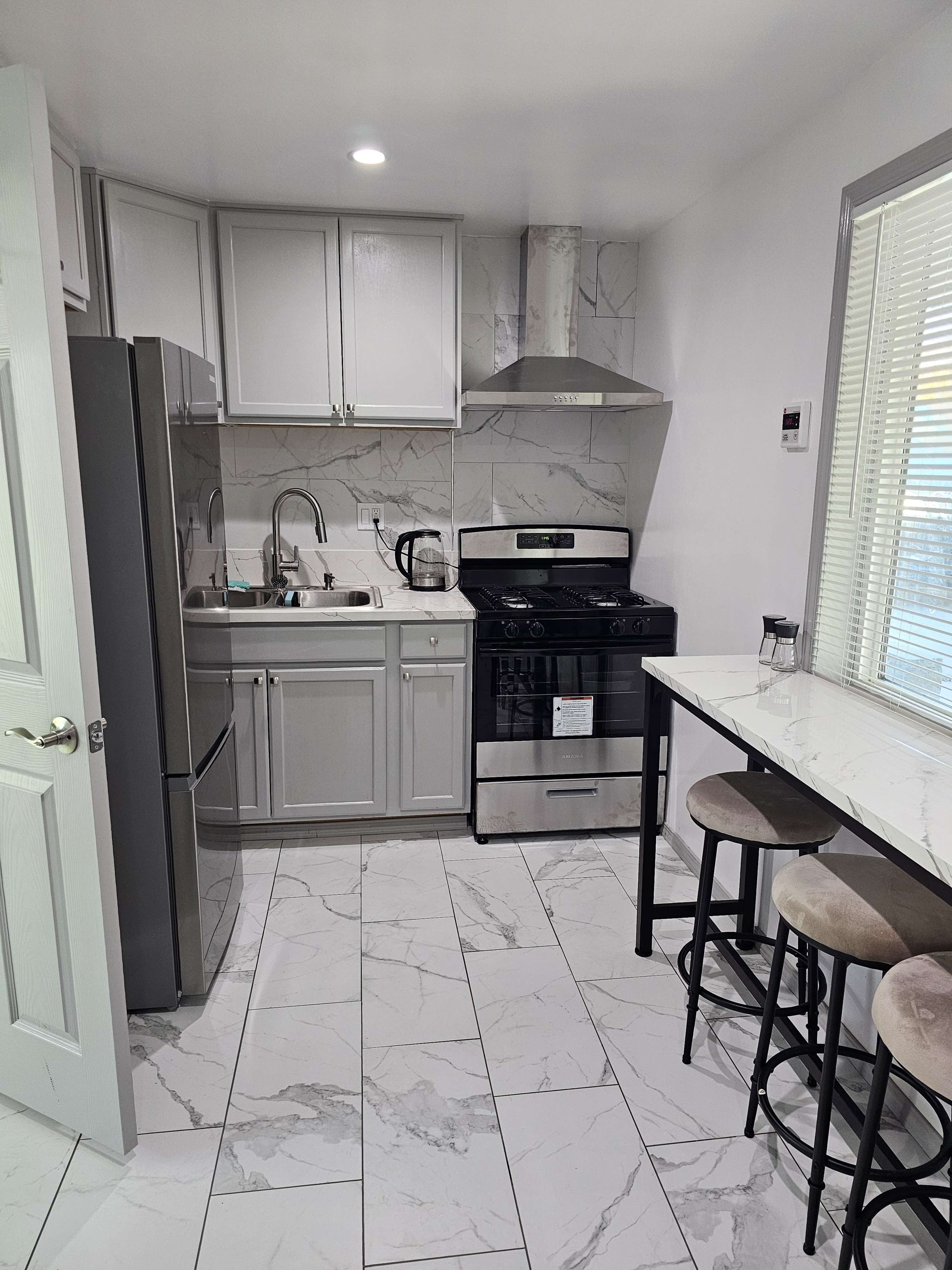 The image shows a modern kitchen featuring gray cabinets, a stainless steel refrigerator, a gas stove, and a white table with black metal stools on a tiled floor.