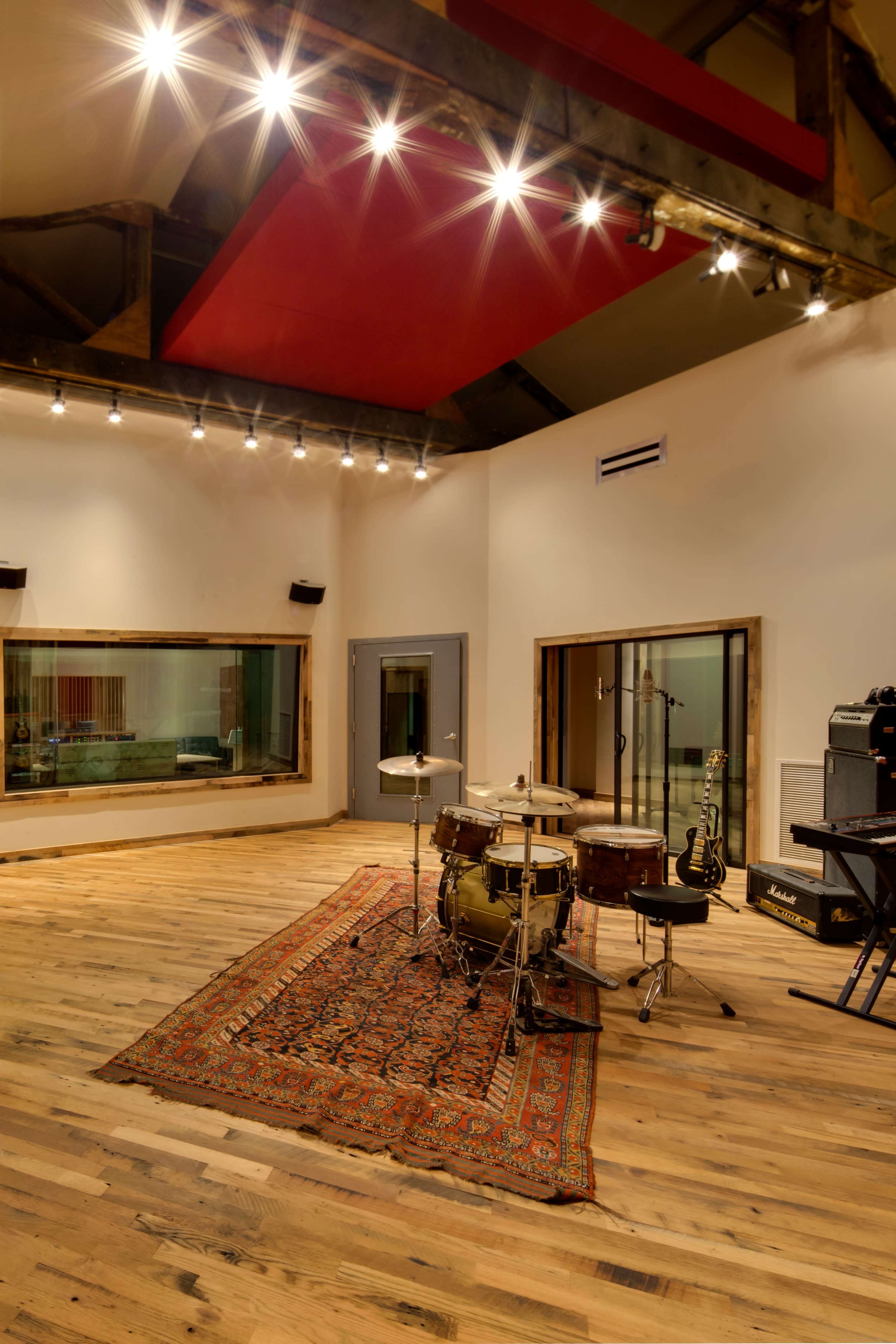 A spacious music studio features a drum set on a large area rug, with glass soundproofing panels and musical instruments in the background.