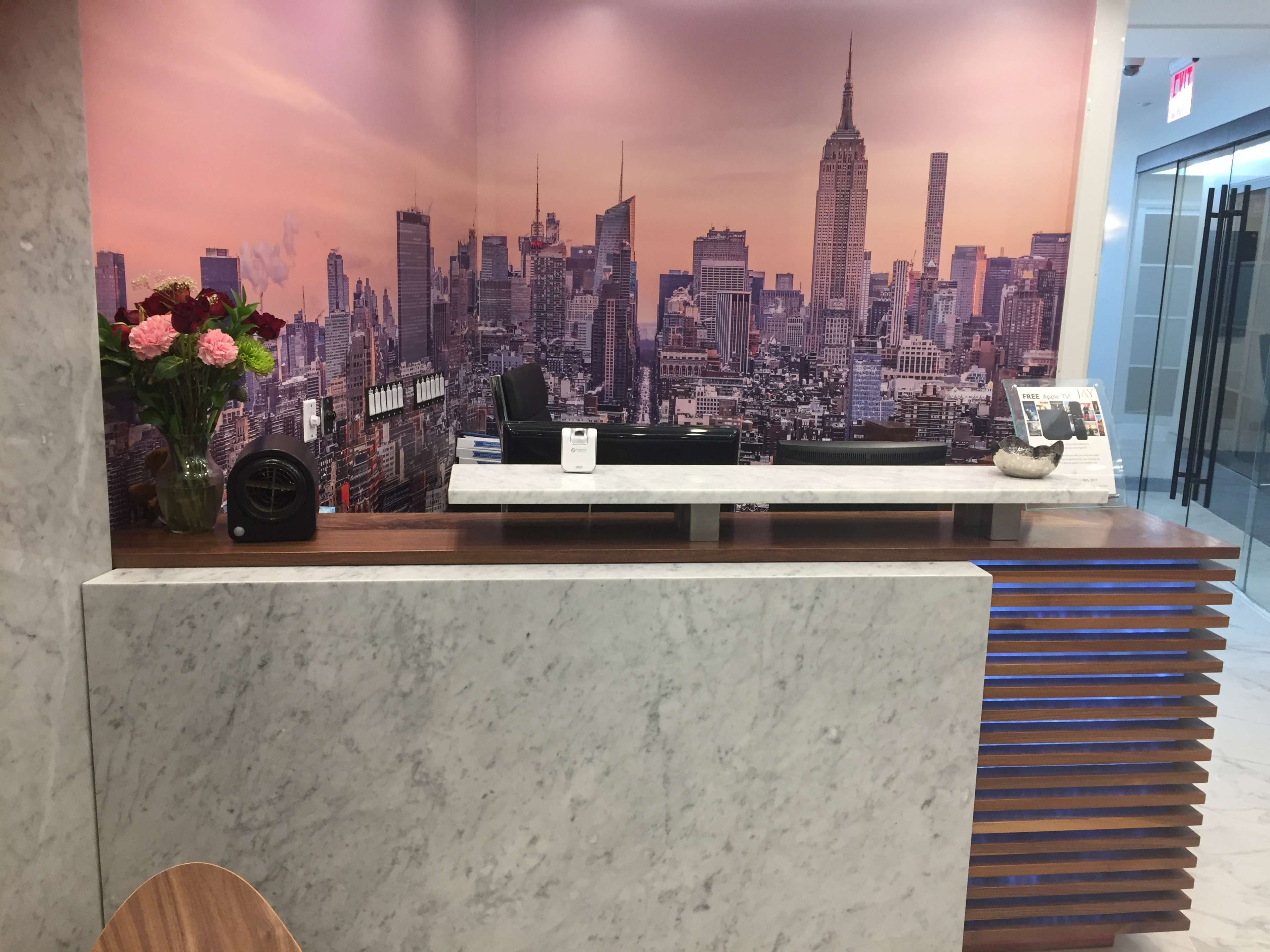 A reception area featuring a marble front desk, a floral arrangement, and a large wall mural of a New York City skyline at sunset.