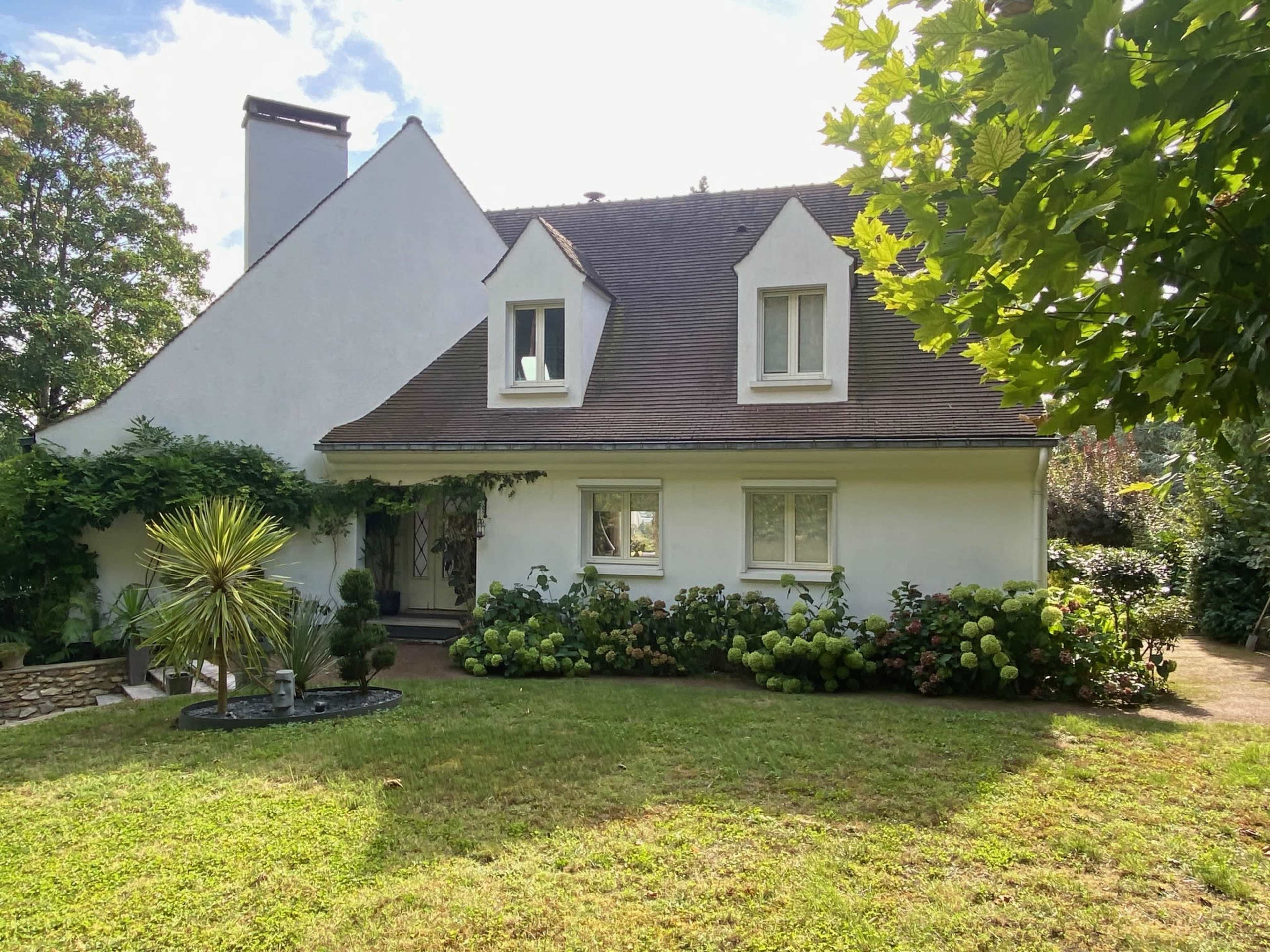 The image shows a white, two-story house with a sloped roof, surrounded by a well-maintained garden featuring green shrubs and blooming hydrangeas.