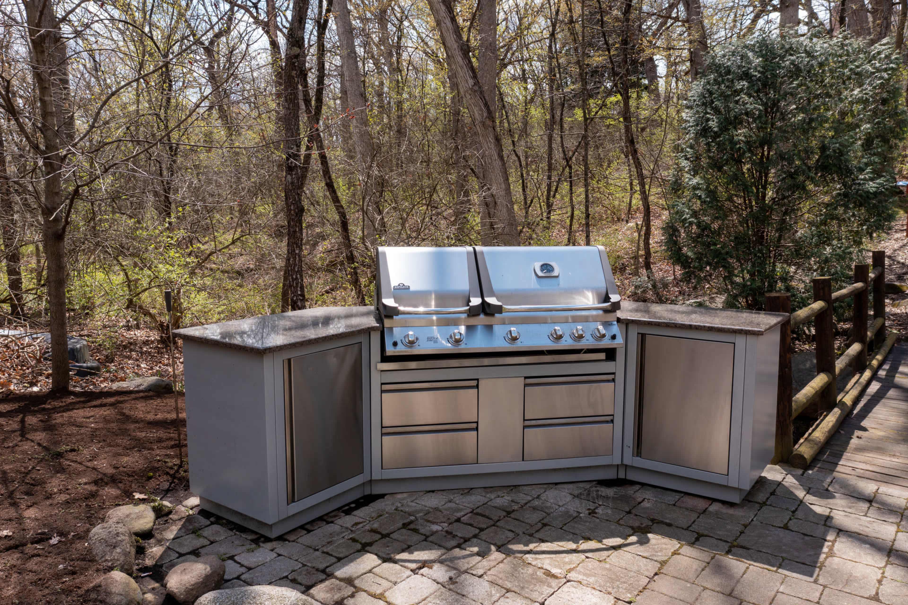 A stainless steel outdoor kitchen grill station set on a stone patio surrounded by trees.