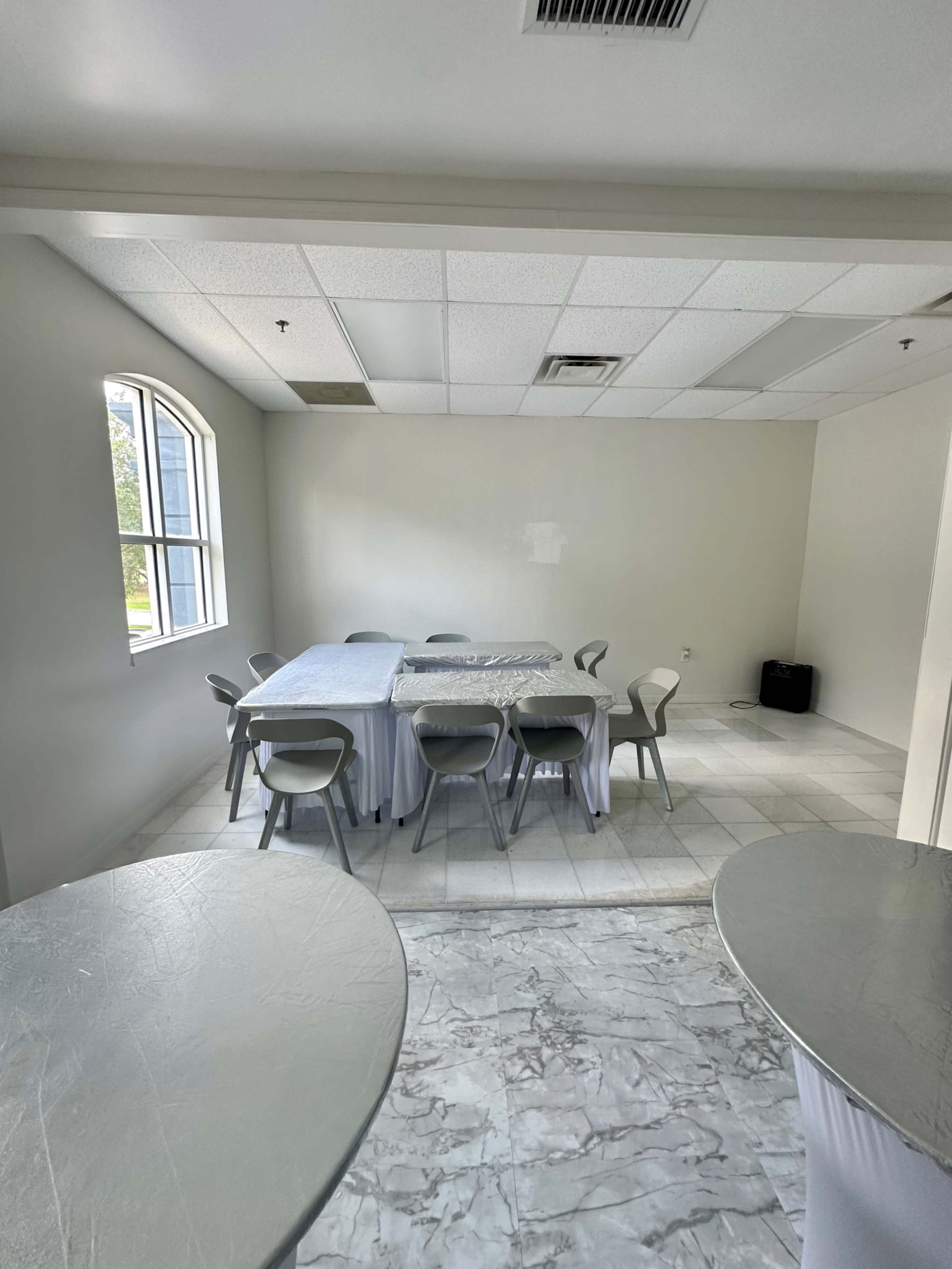 A sparsely furnished room features a table surrounded by chairs, with two small round tables in the foreground and a large window letting in natural light.