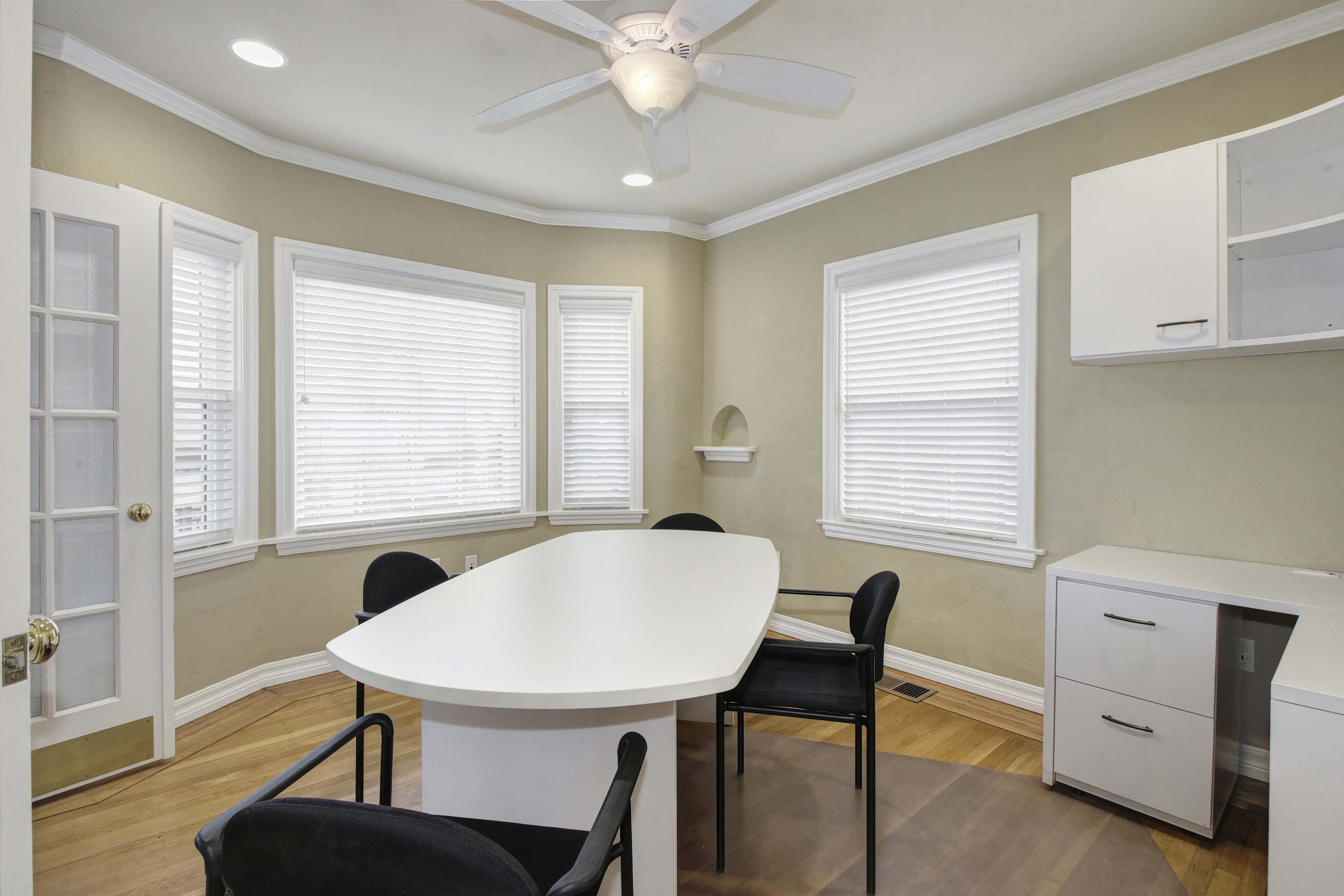 A brightly lit room features a large white table surrounded by black chairs, with multiple windows allowing natural light to enter.