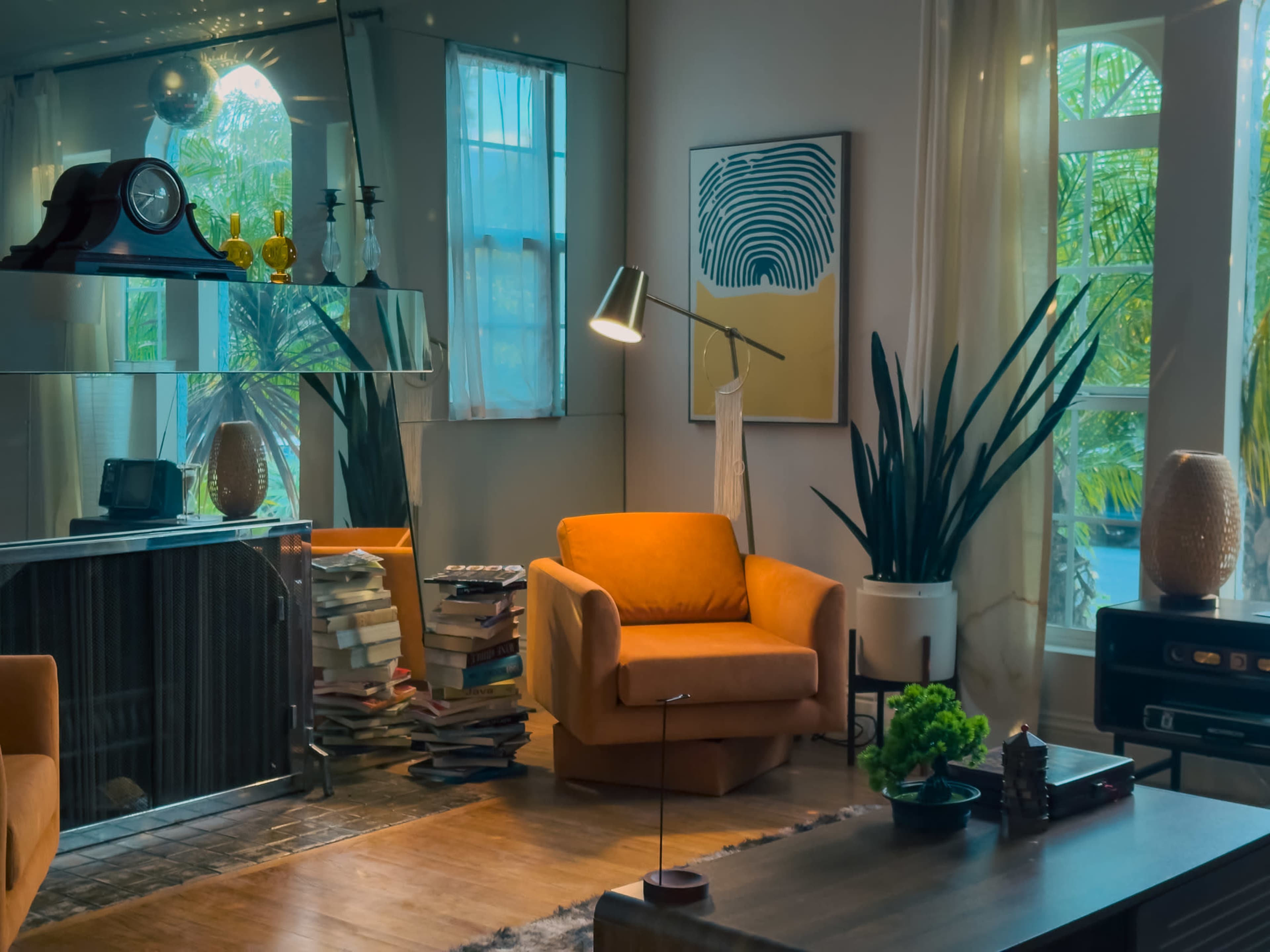 The image shows a modern living room featuring an orange armchair, a stack of books, plants, and a large mirror reflecting a window.