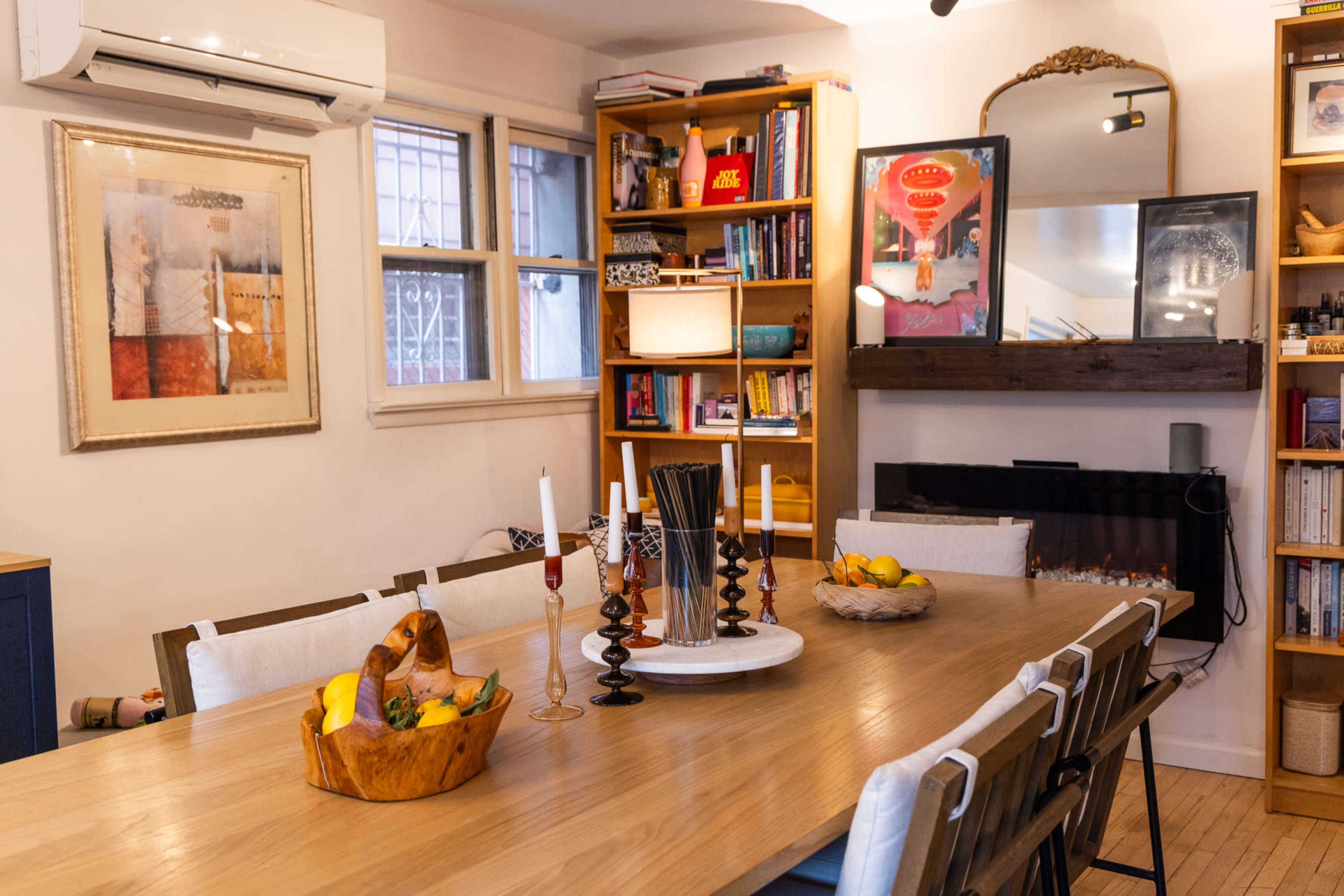 The image shows a cozy dining area with a wooden table surrounded by chairs, a lit fireplace on one side, and bookshelves filled with various items and decorations.