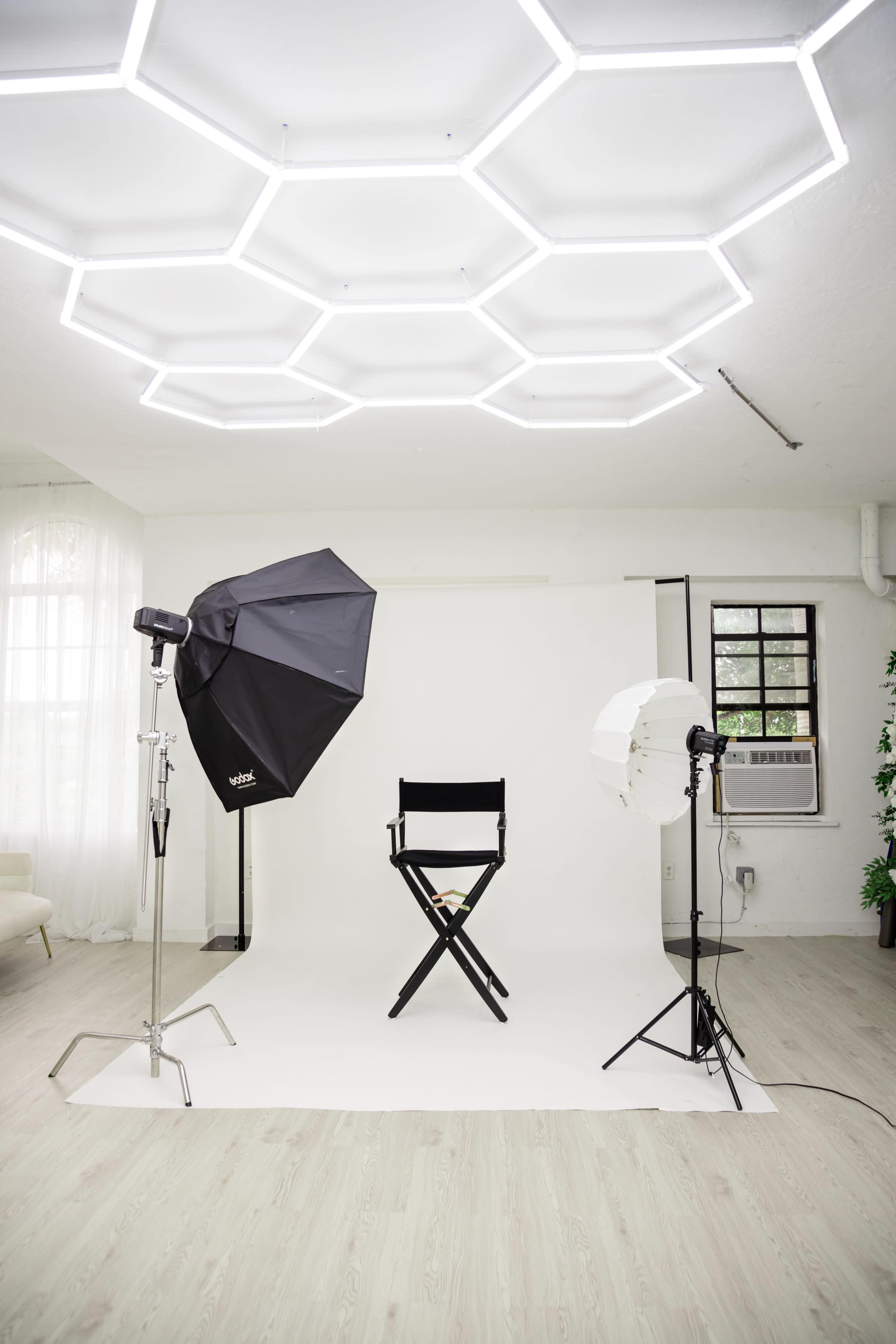 The image shows a minimalist photography studio with a white backdrop, a director's chair, and two lighting setups positioned to either side.