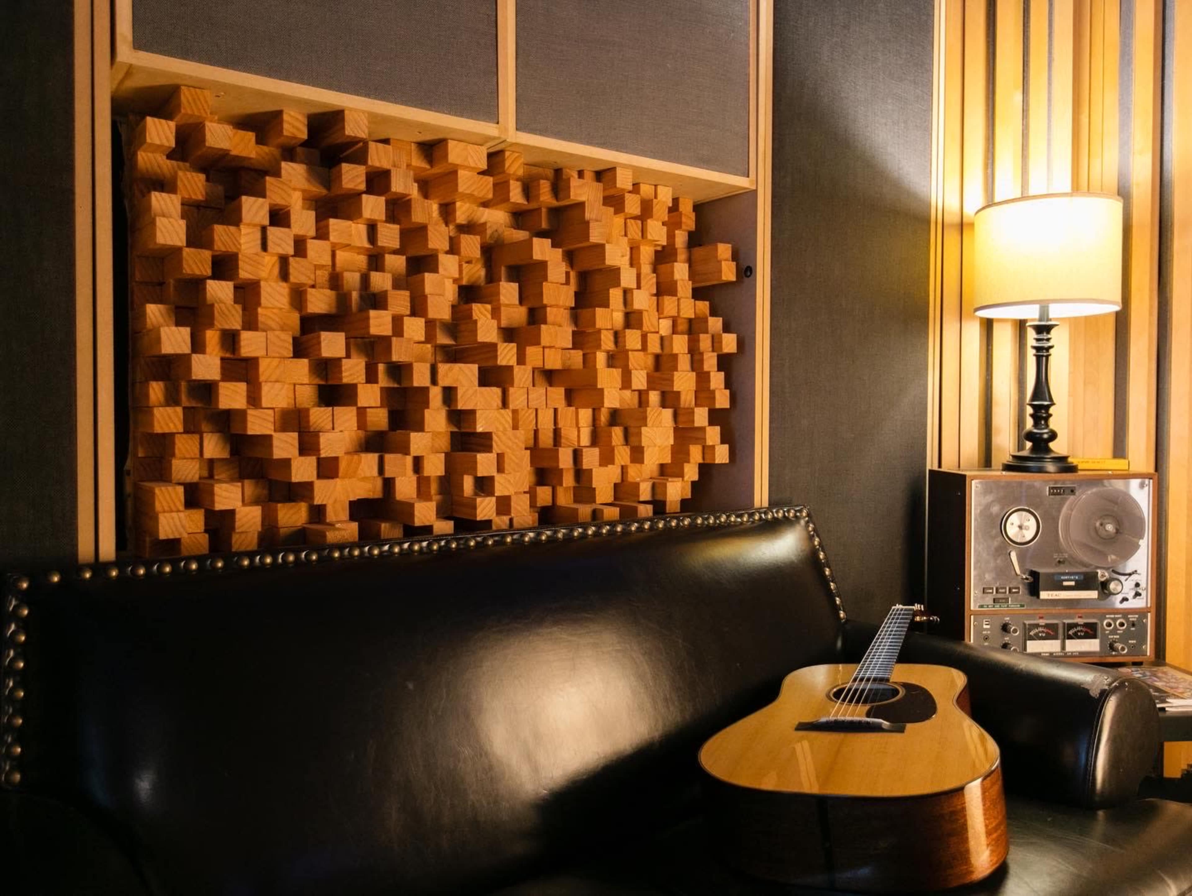 A wooden wall panel made of small blocks is behind a black leather couch with an acoustic guitar resting on it, next to a lamp and a vintage audio equipment setup.
