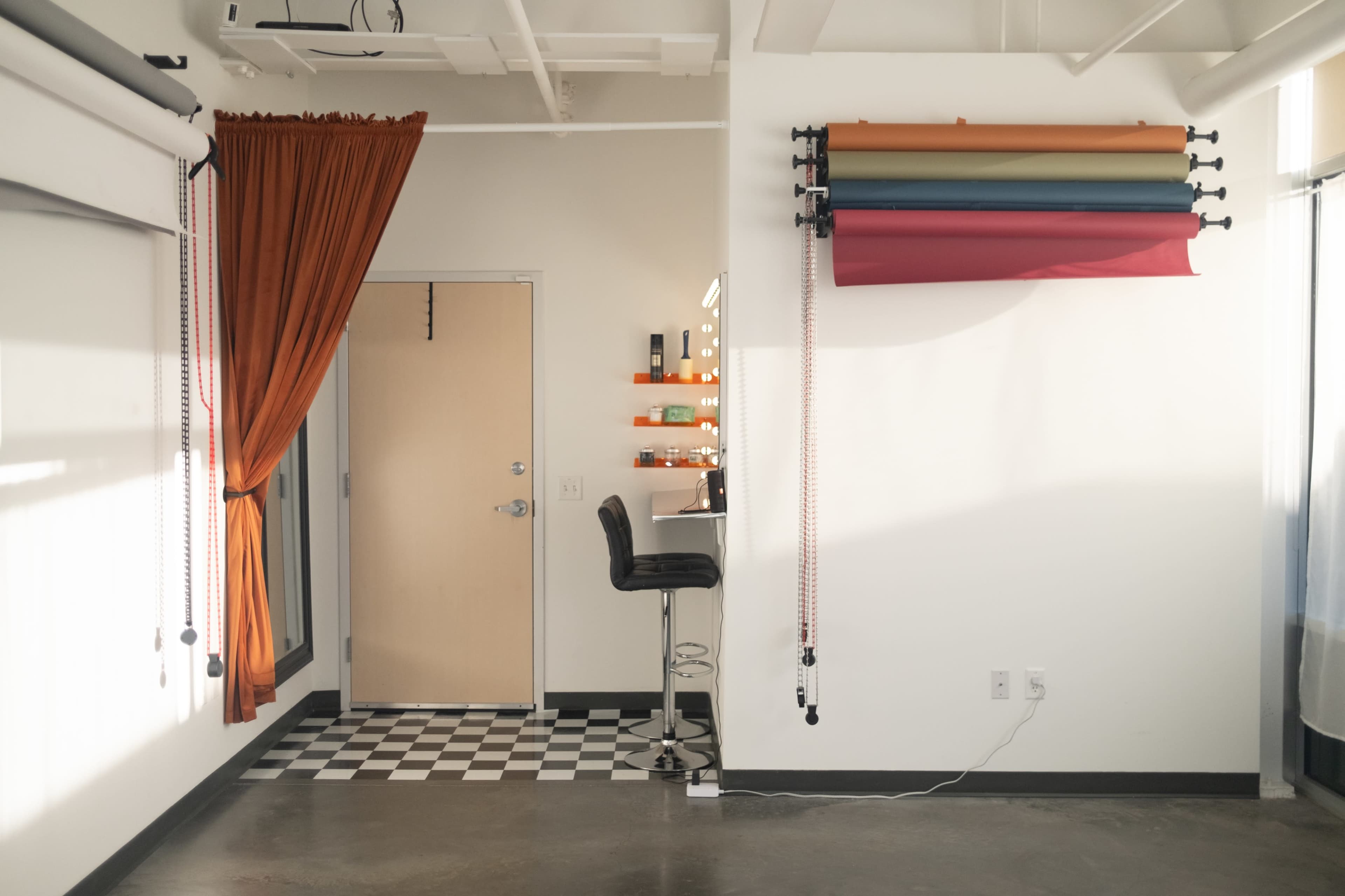 The image shows a contemporary studio space with a barstool near a checkered floor and colorful backdrop rolls mounted on the wall.