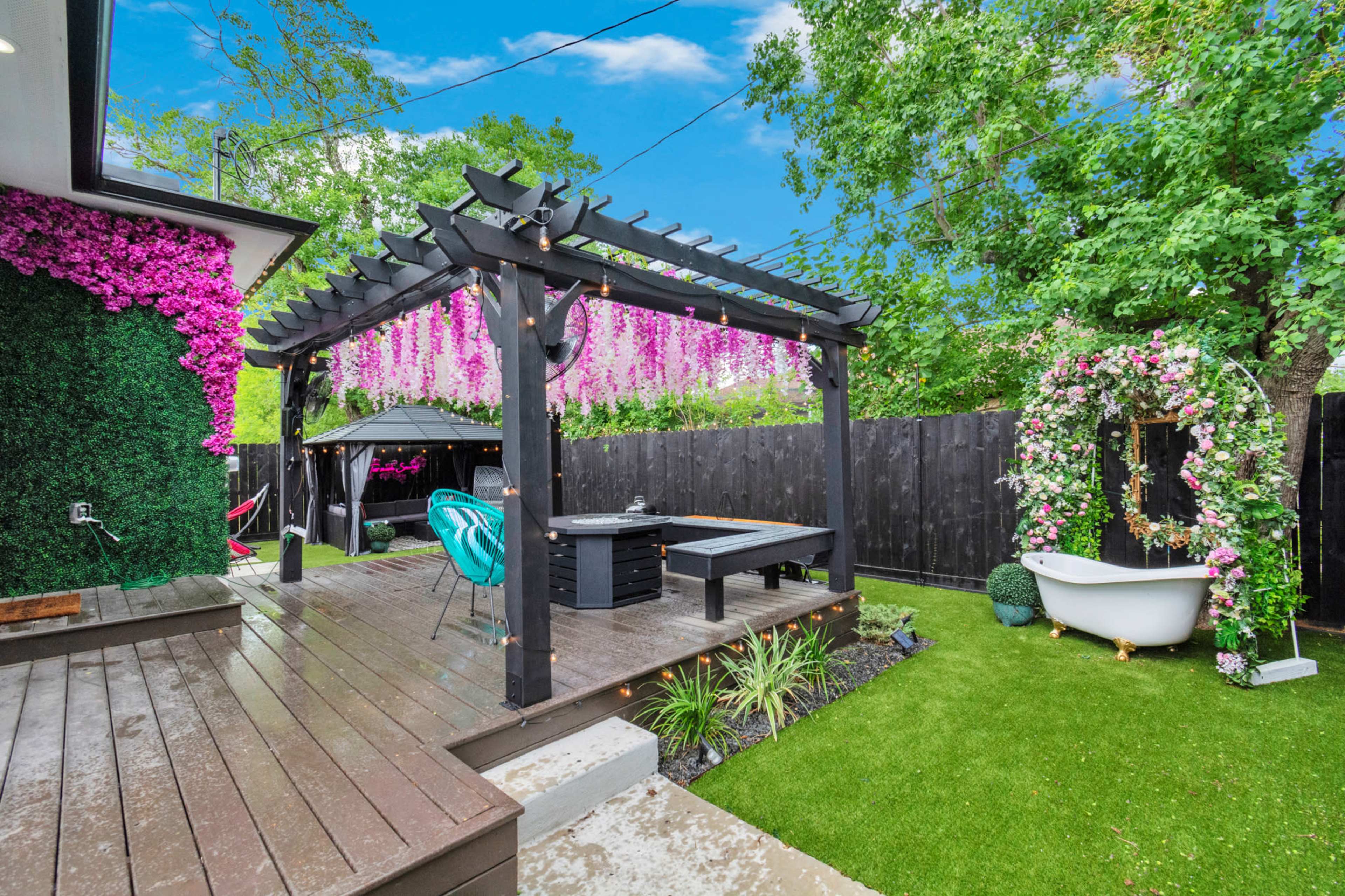 A backyard scene with a wooden deck, a pergola adorned with hanging flowers, a gazebo, and a freestanding bathtub surrounded by artificial grass.