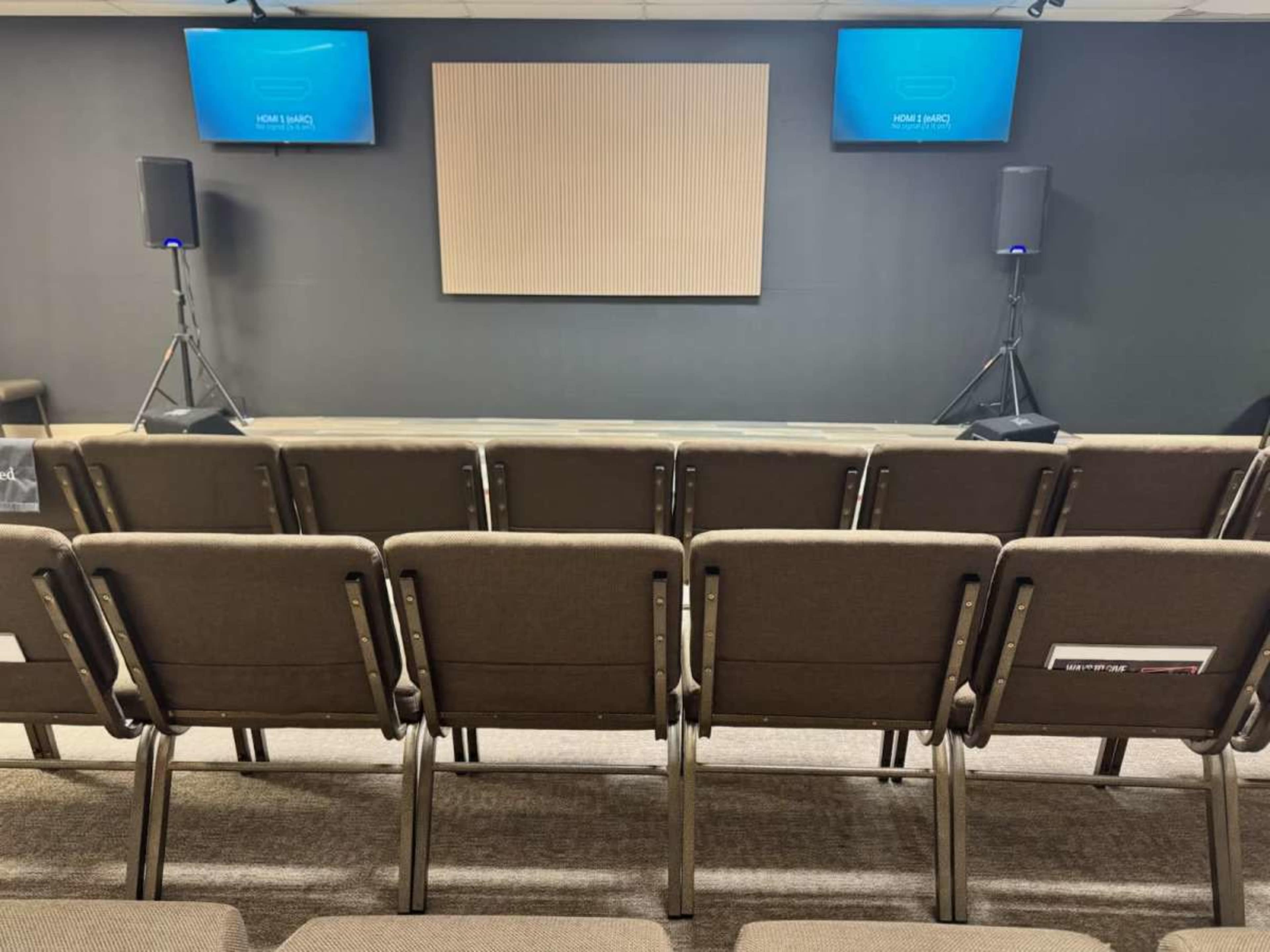The image shows a conference room setup with several rows of chairs facing a blank wall and two speakers on either side.