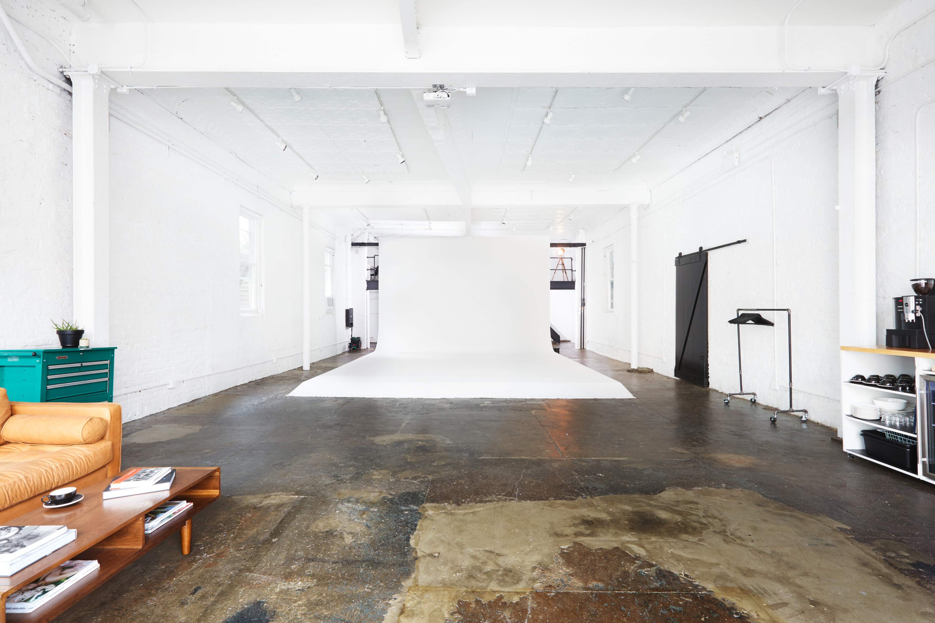 The image shows a spacious studio with white walls, a large empty backdrop, and a seating area featuring an orange couch and wooden coffee table.