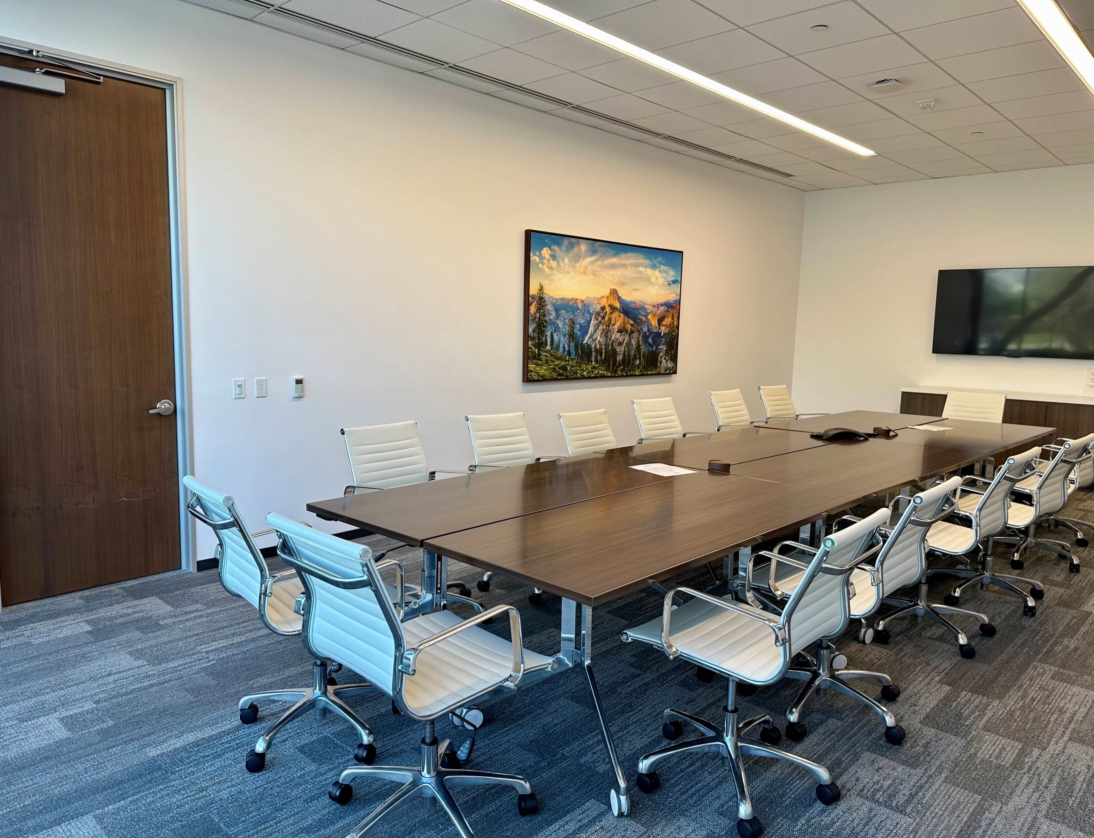 A large conference room features a long wooden table surrounded by white chairs, with a landscape painting on the wall and a screen on one side.