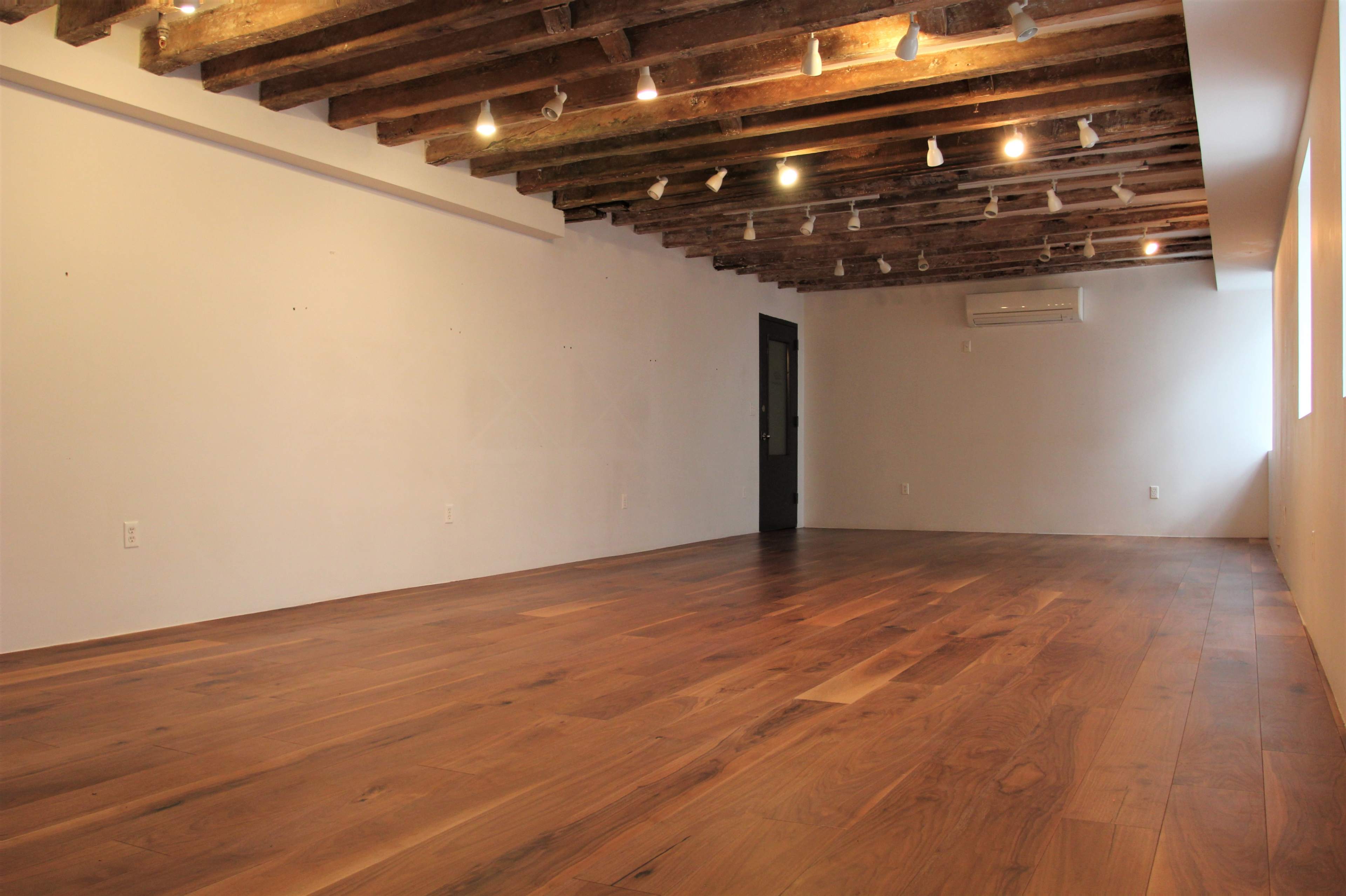 An empty room with wooden flooring, exposed ceiling beams, and track lighting along the ceiling.